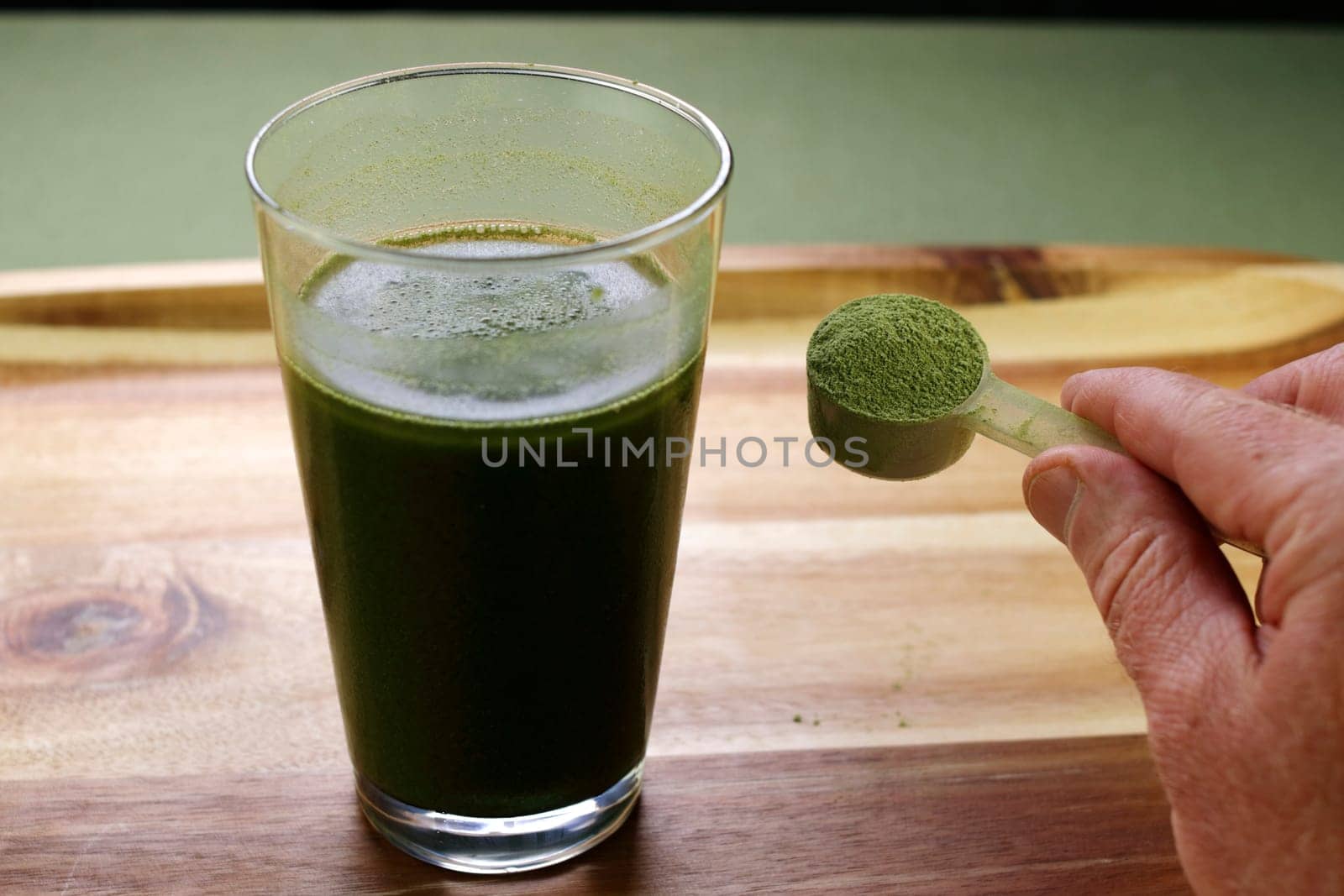 A scoop of organic celery powder being shown by fingers of a white hand near a celery powder and aloe vera juice beverage. Aloe vera juice with celery powder drink near a hand showing celery powder.