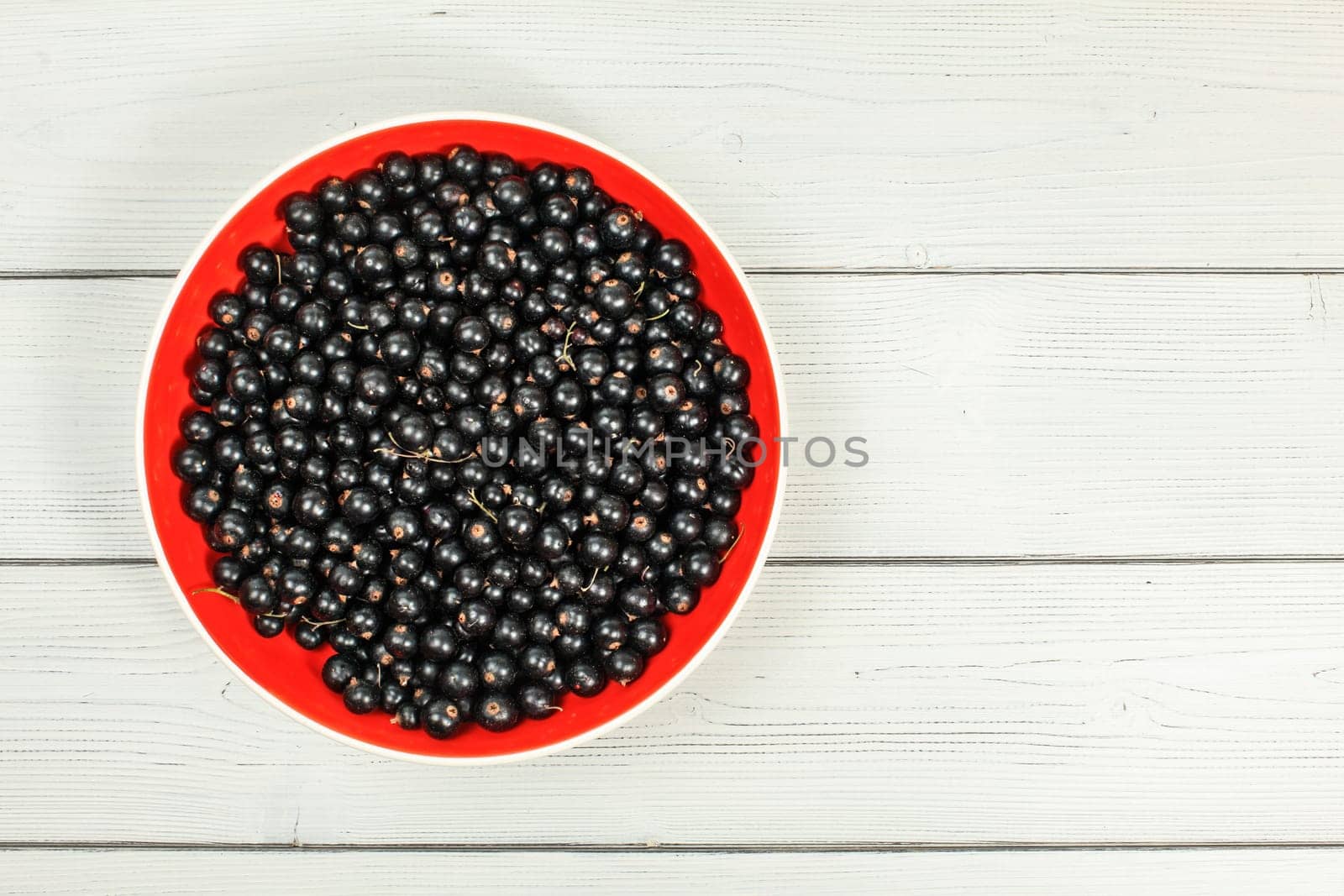 Tabletop view, red ceramic bowl full of freshly harvested blackcurrants, on white boards desk.