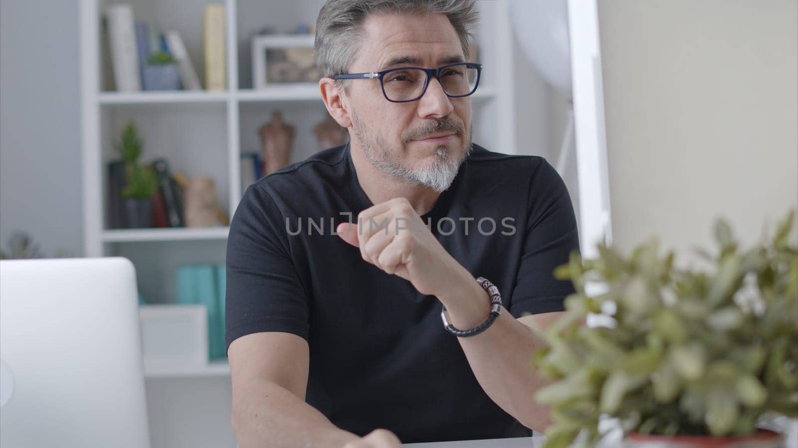 Older white man in glasses sitting at desk using desktop computer in home office. 50s businessman staying home in casual working online thinking on business solution, smiling.