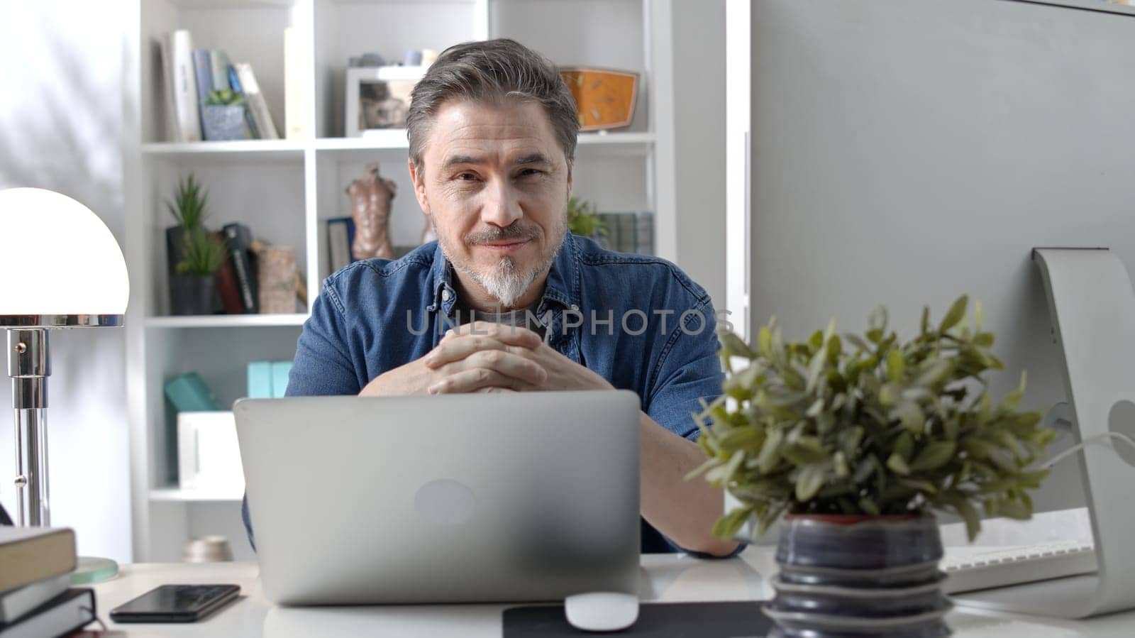 Portrait of happy older man sitting at desk working with laptop in home office. Confident 50s businessman in casual with gray hair smiling.