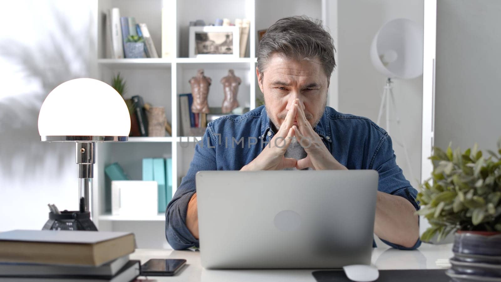 Older white man in glasses sitting at desk using laptop in home office. 50s businessman staying home in casual working online with computer thinking on business solution.