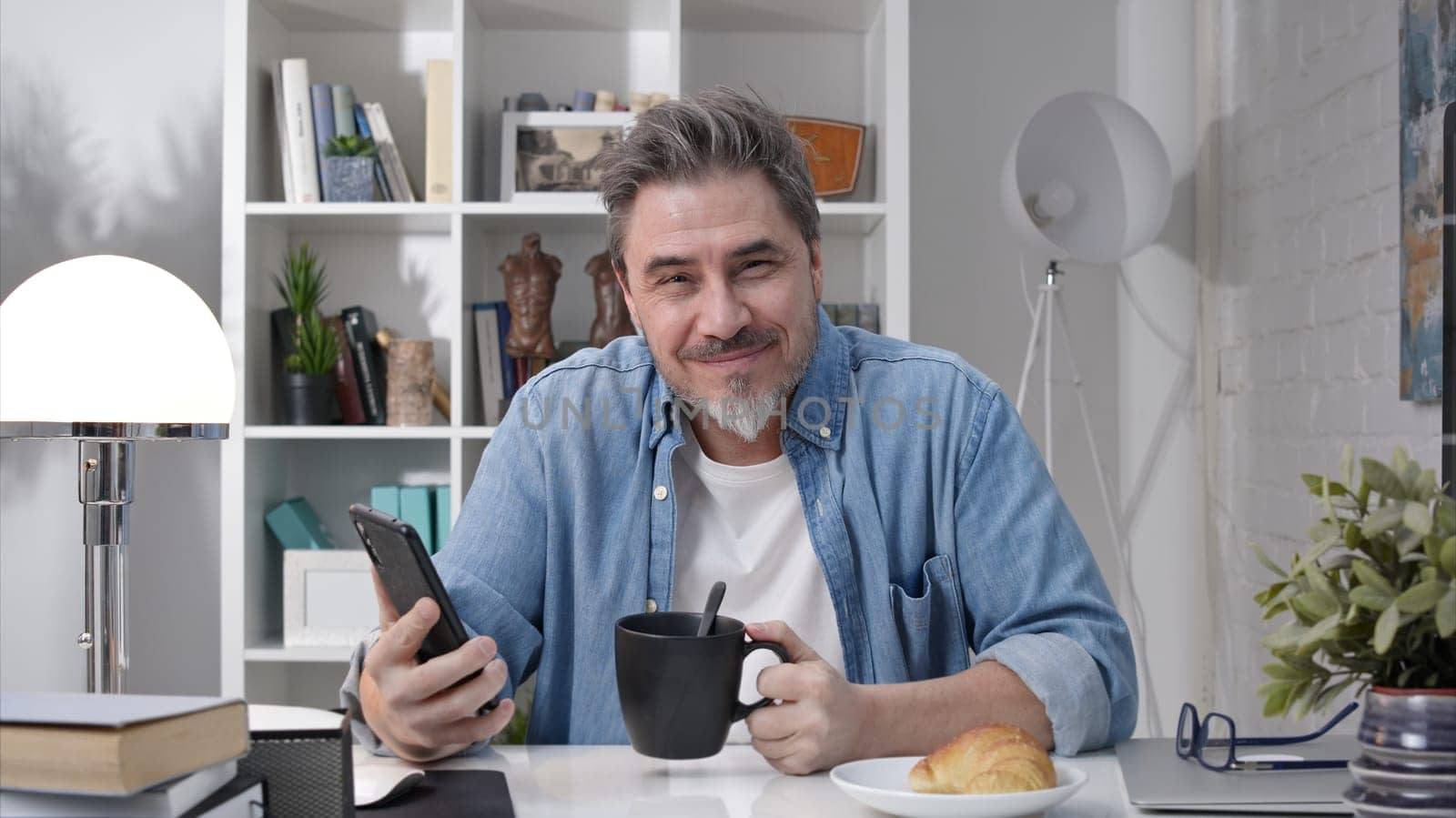 50s man having breakfast checking news on phone drinking morning coffee at home. Confident happy smiling.