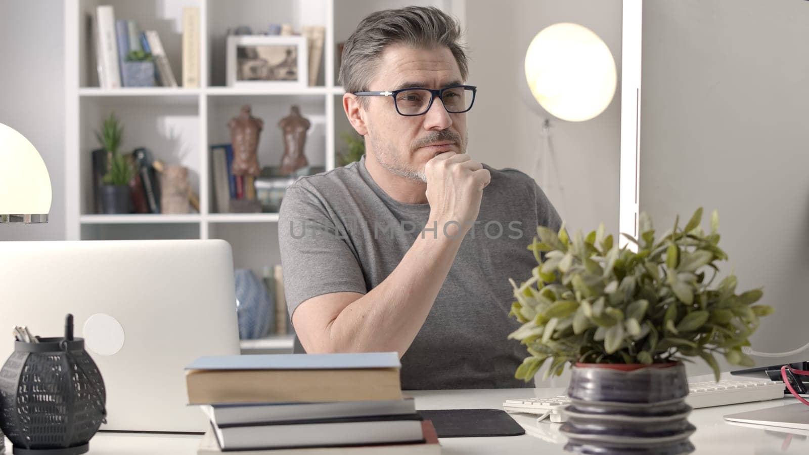Older white man in glasses sitting at desk using laptop in home office. 50s businessman staying home in casual working online with computer thinking on business solution.