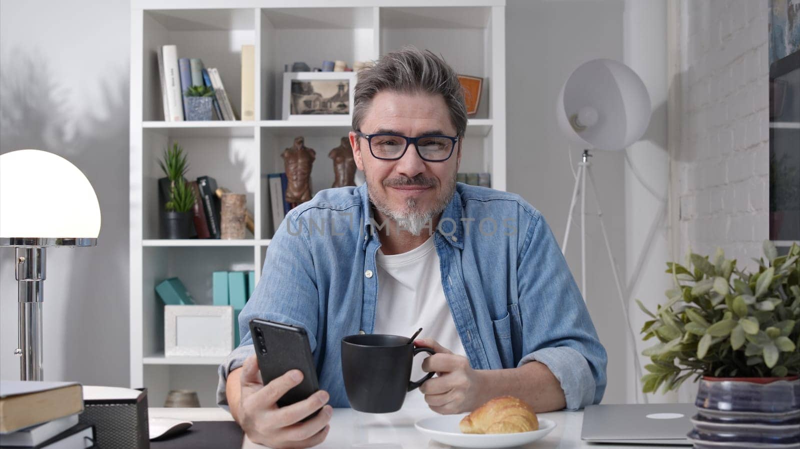 50s man having breakfast checking news on phone drinking morning coffee at home. Confident happy smiling.