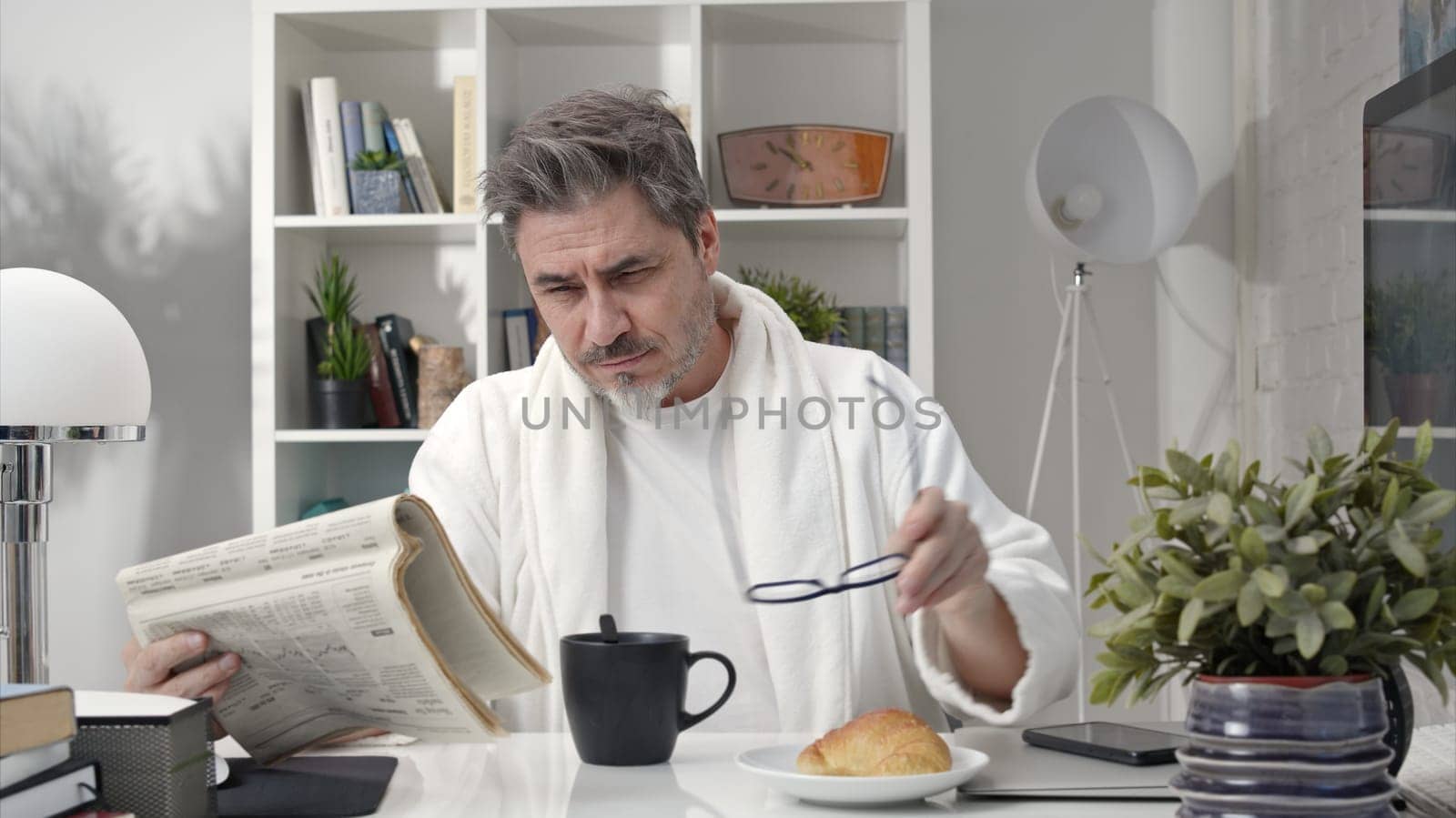 50s man having breakfast drinking morning coffee at home. Businessman checking newspaper for business and stock financial news.