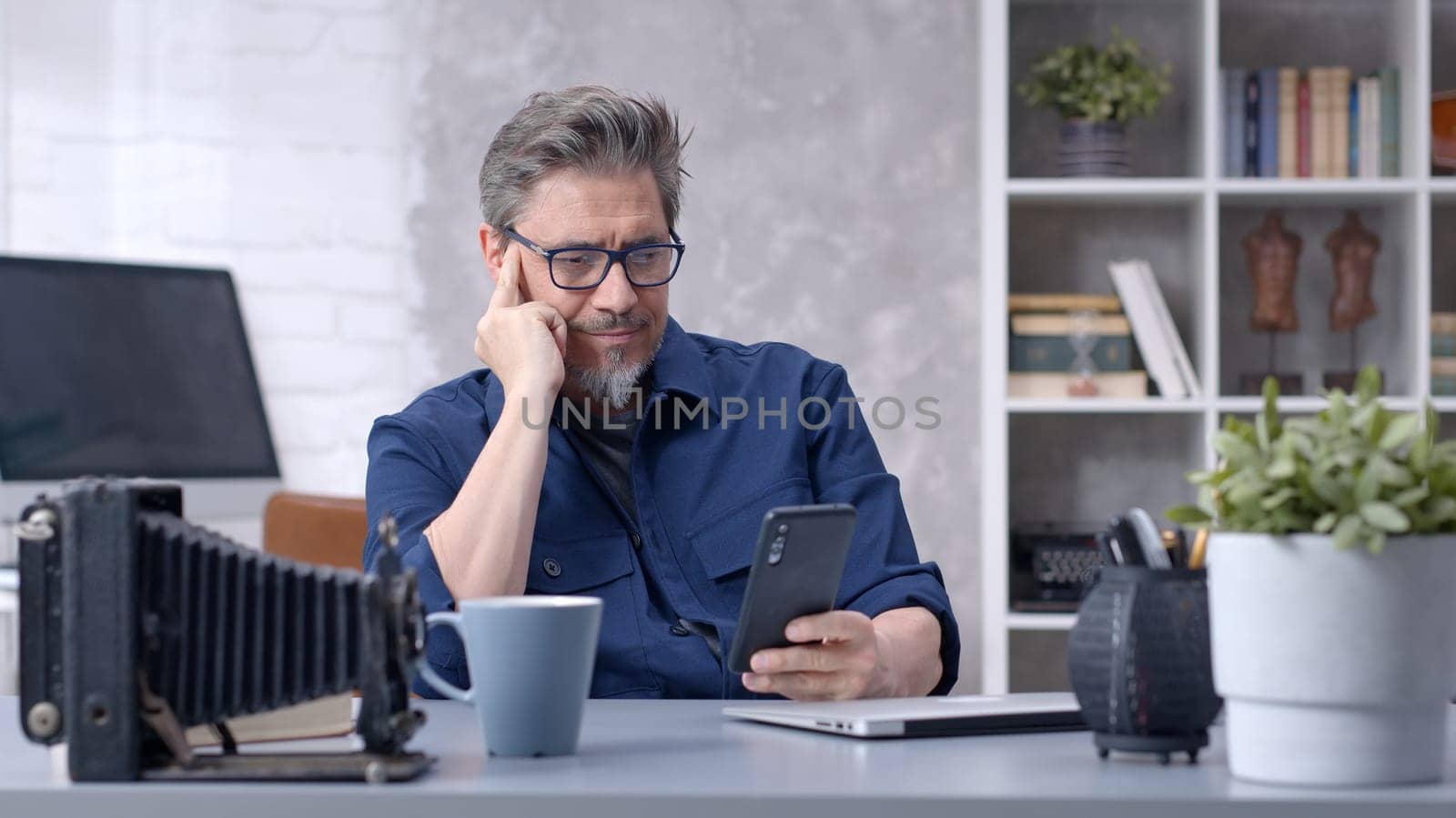 Bearded man working online with laptop computer and smart phone at home sitting at desk. Businessman in home office. Portrait of mature age, middle age, mid adult man in 50s.