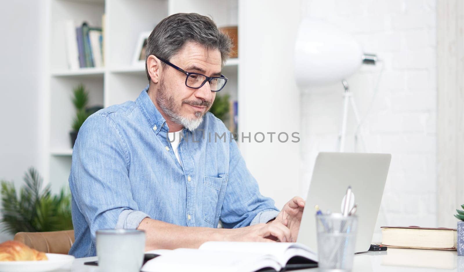 Older man working online with laptop computer at home sitting at desk. Home office, browsing internet, study room. Portrait of mature age, middle age, mid adult man in 50s.