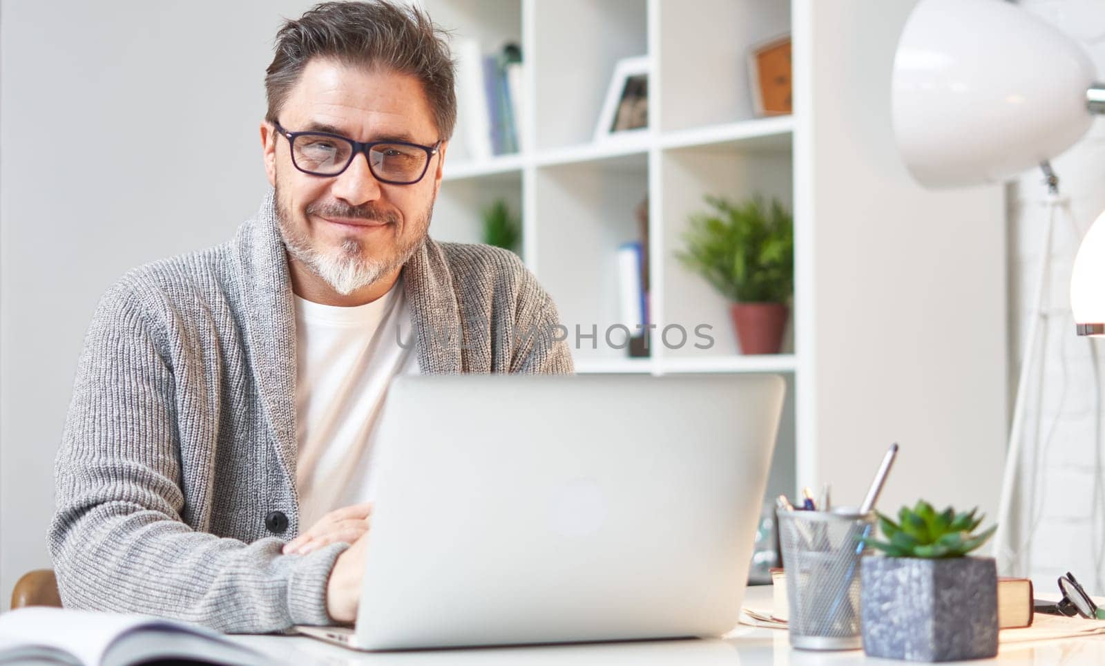 Happy older man working online with laptop computer at home sitting at desk. Home office, browsing internet, study room. Portrait of mature age, middle age, mid adult man in 50s.