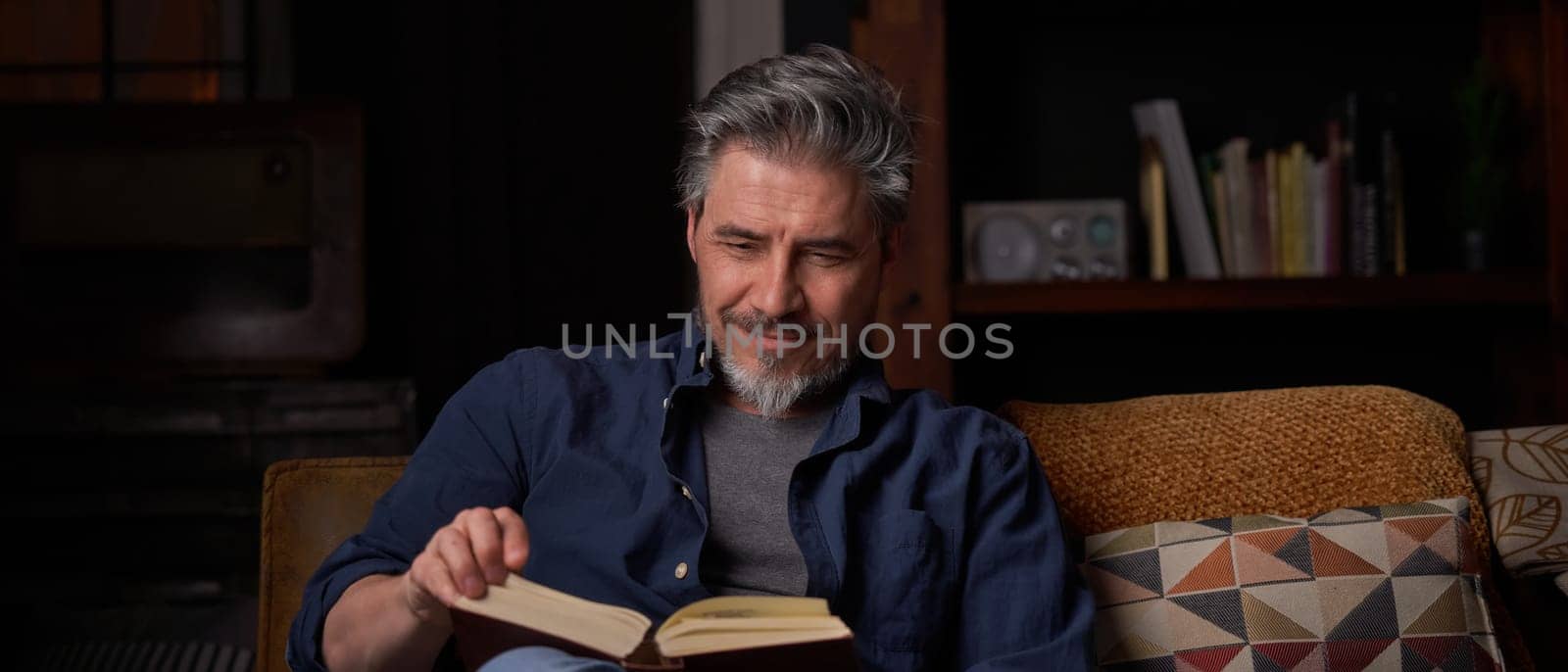 Happy man sitting on couch at home in cosy room reading book. Relaxing, leisure, education. ..Portrait of mature age, middle age, mid adult man, bearded, smiling, authentic look.