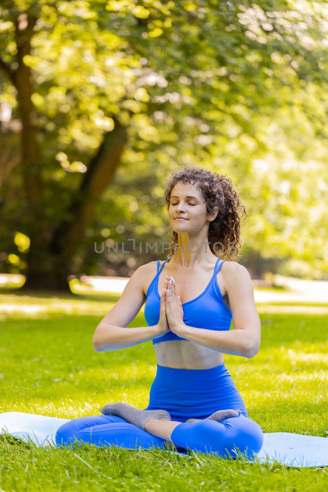Young woman sitting on mat in lotus position, relaxing in park on summer day. Sporty carefree adult girl practicing yoga meditation, receives positive energy, healthy lifestyle. Active sportswoman.