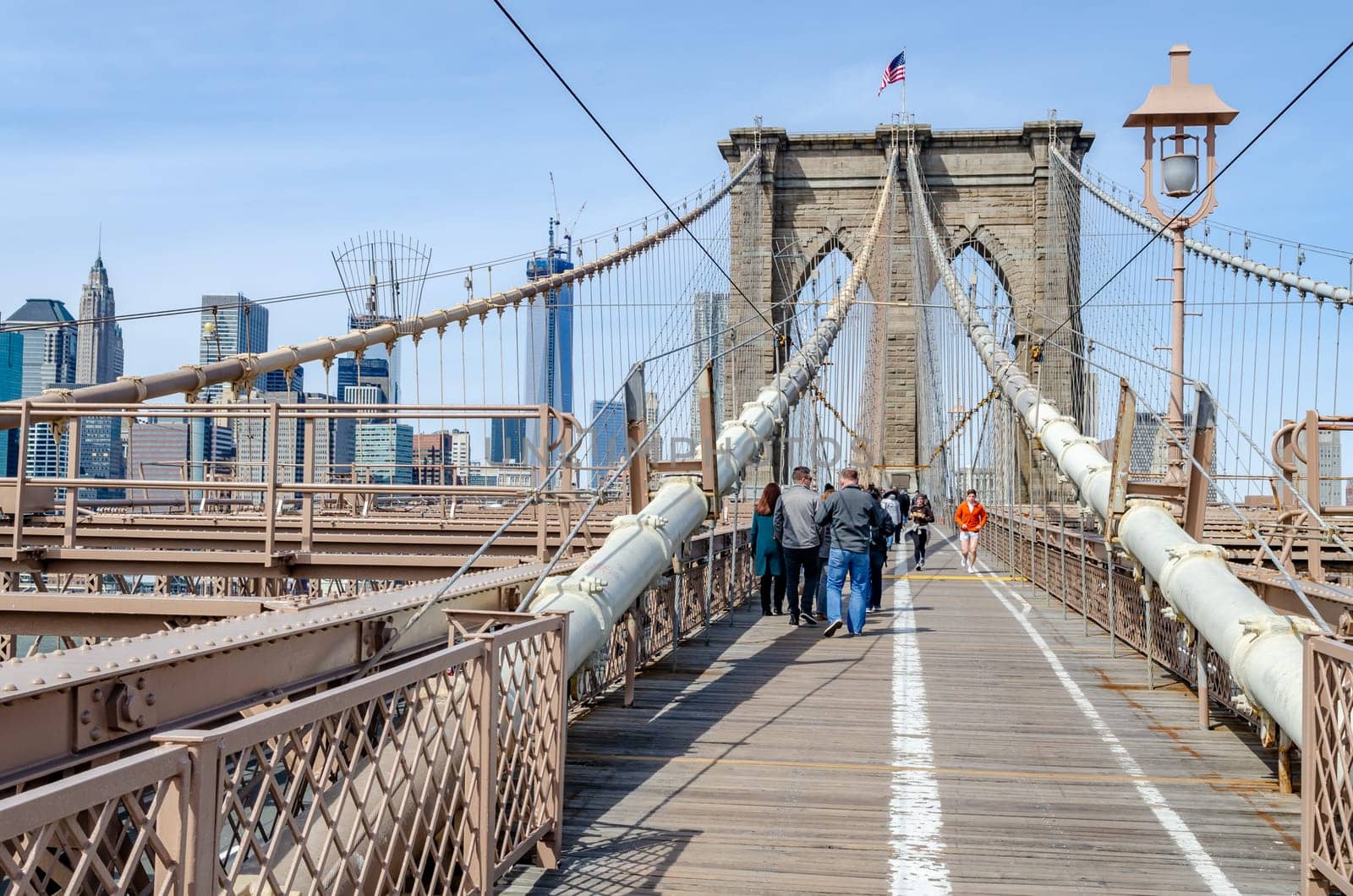 Two People jogging on the right side of the Brooklyn Bridge New York City during daytime, lots of tourist walking on the left side of the Bridge, wire ropes and street lamp in the forefront, skyline of Manhattan in the Background, Horizontal