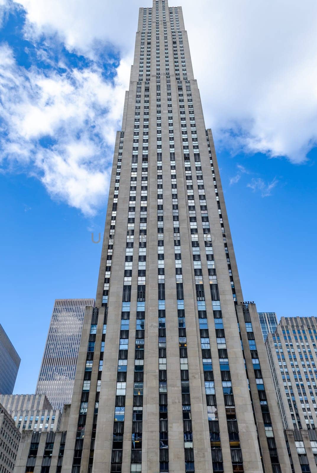 Rockefeller Center building facade, New York City, low angle view during daytime with blue sky and some clouds, reflection on the windows of the building, vertical