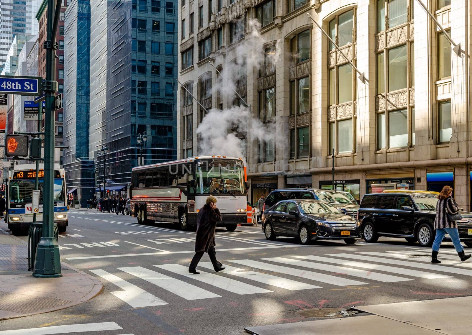People crossing the Street on a crosswalk in Manhattan at East 48th Street, Cars and two busses waiting in the background, Smoke rises on the street behind a bus,  New York City during daytime, horizontal