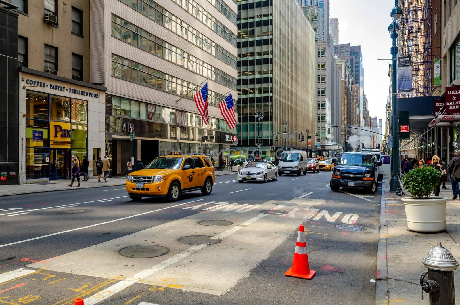 Manhattan, New York City, Street with lots of traffic, Yellow Taxi Cabs and other private cars, People walking on the sidewalk, Coffee Pastry Prepared Food Store in the background, American Flags hanging down a Building, horizontal