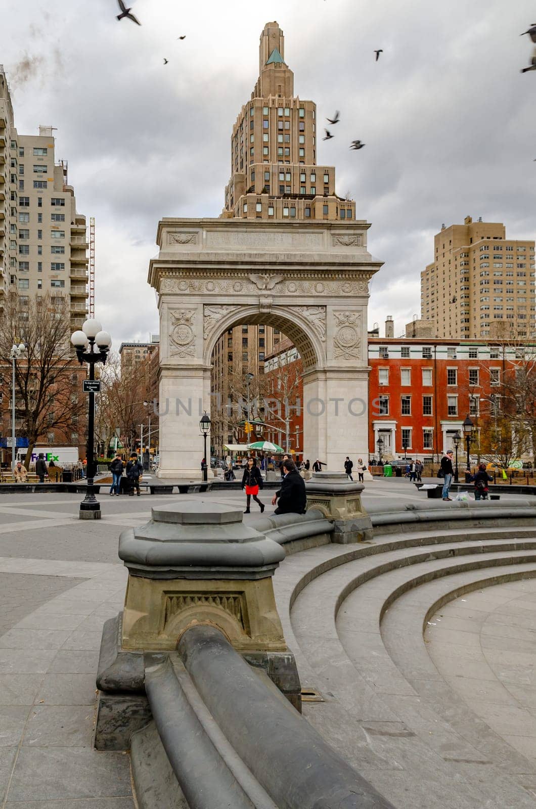 Washington Square Arch, New York City during a cold winter day with empty fountain in forefront, Birds flying over the building, people walking in the park, vertical
