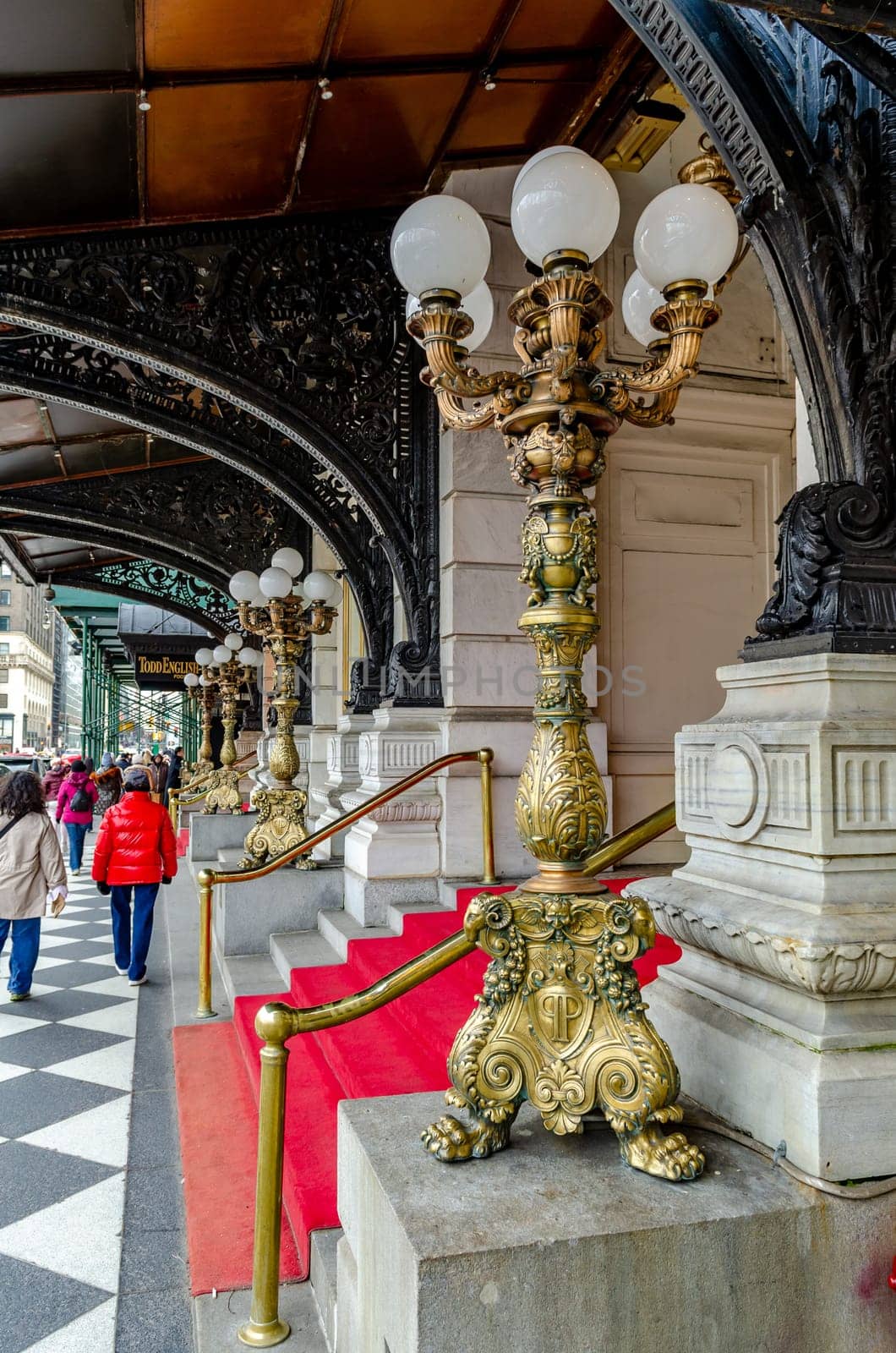 Entrance of The Plaza Hotel, New York City with golden Street Lamps and a red carpet in front of the Building, View from the side, People walking on the sidewalk in the background, vertical