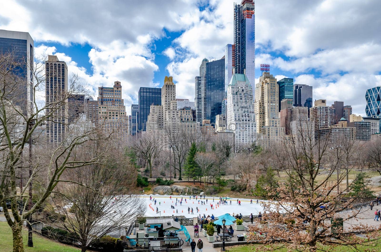 Trumps Wollman Rink with People Ice Skating during daytime in winter, view from the distance, Skyline of Manhattan in the Background, Central Park New York City, benches and people walking in the forefront, horizontal