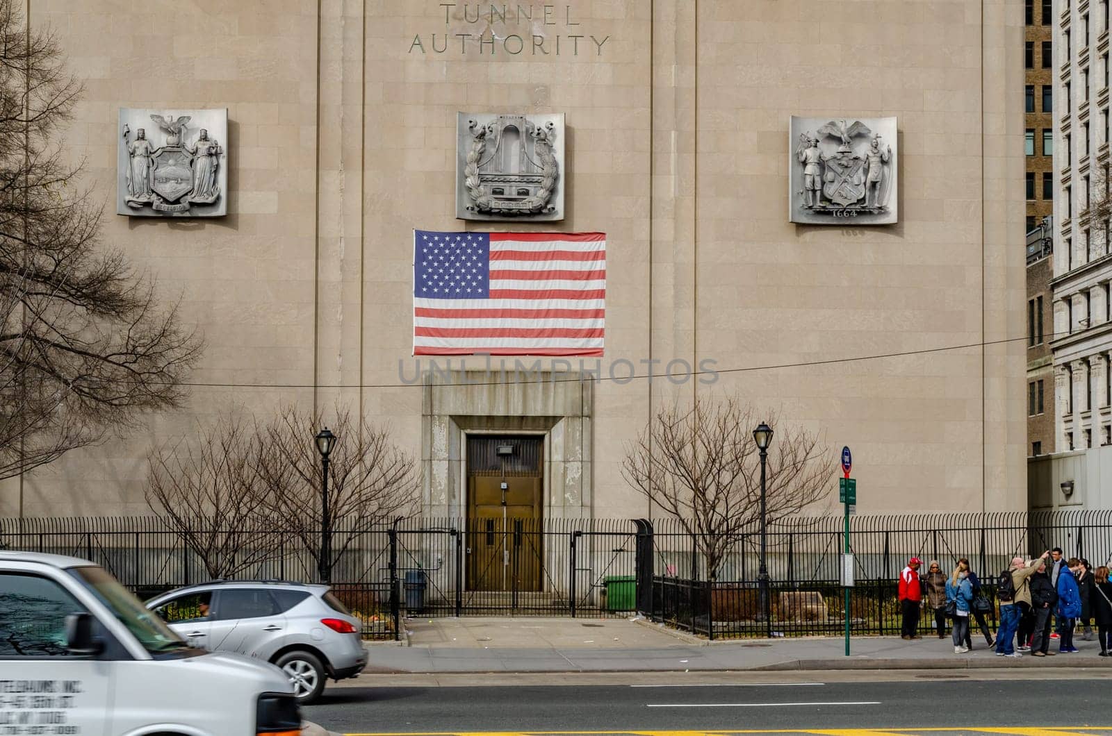 New York City Triborough Bridge and Tunnel Authority Building with Building Entrance and American flag on top of it, street with traffic and walking people in forefront, during winter, horizontal