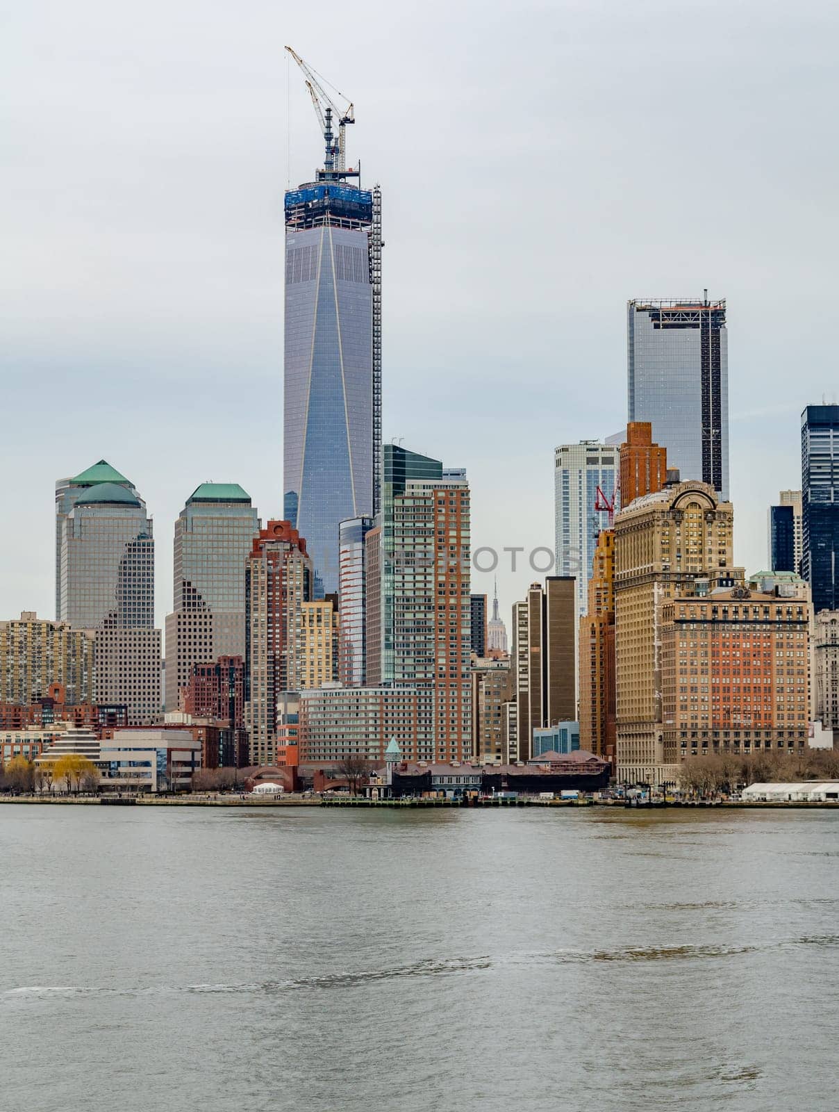 One World Trade Center with construction area and Crane on top of it, Manhattan Skyline with Hudson river in forefront, during winter day with overcast, vertical