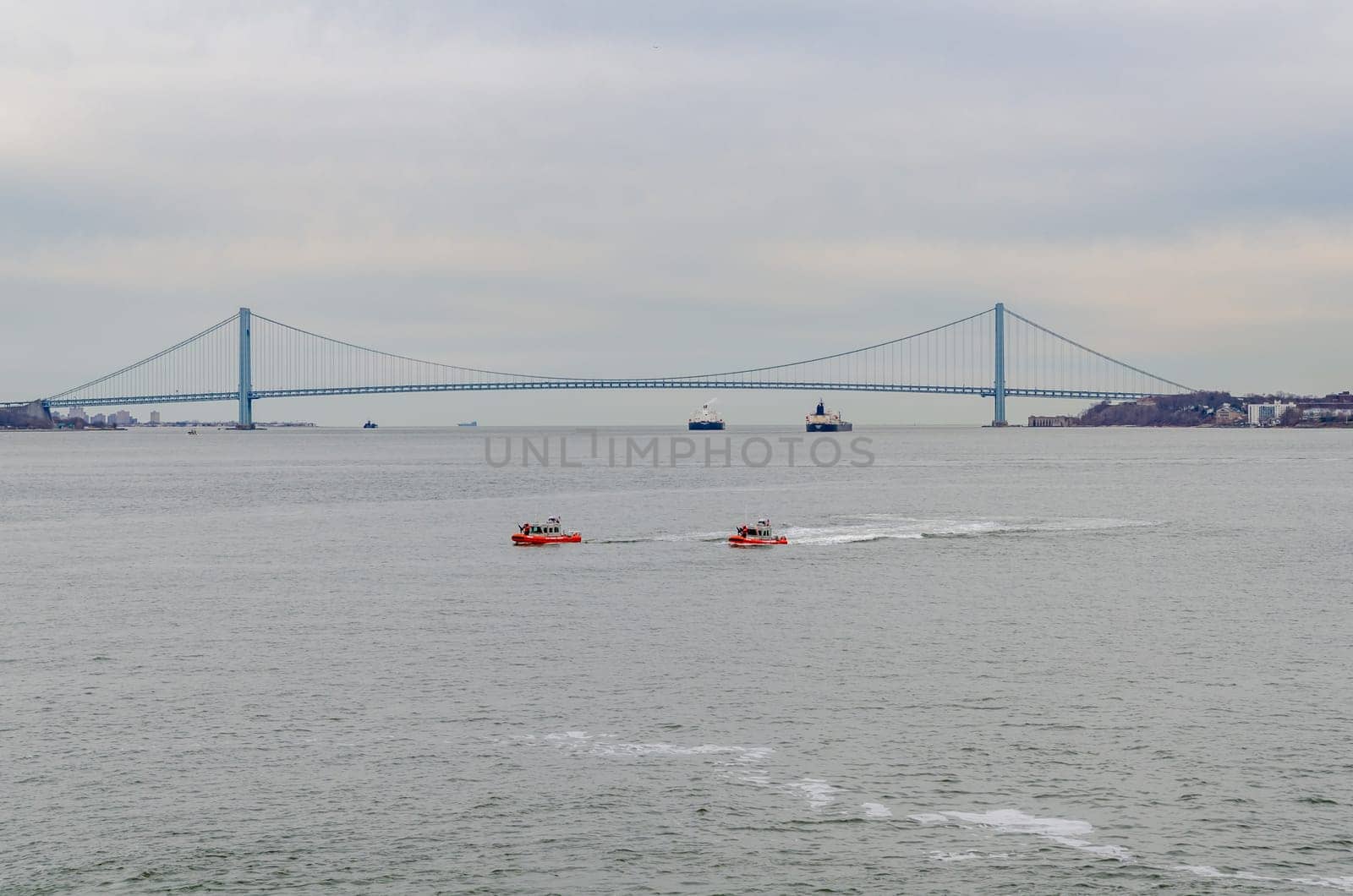 Two red United States Coast Guard ships and two blue and white industrial ships on Hudson river in front of Verrazano-Narrows Bridge, New York City, during winter evening with overcast, horizontal