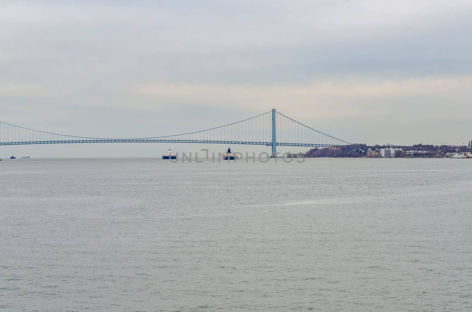 Two industrial ships underneath blue Verrazano-Narrows Bridge, New York City, at the horizon, during winter evening with overcast, horizontal