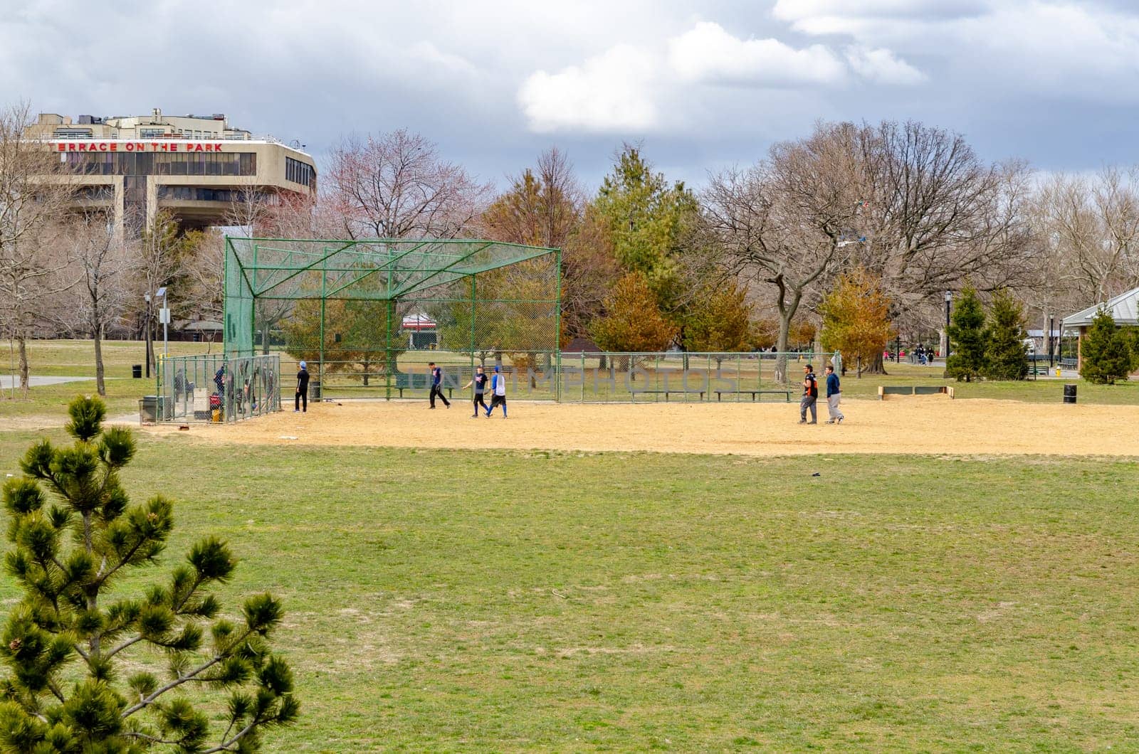 Young male Students playing Baseball in Flushing-Meadows-Park during overcast winter day with Terrace in the Park Restaurant in the background, horizontal