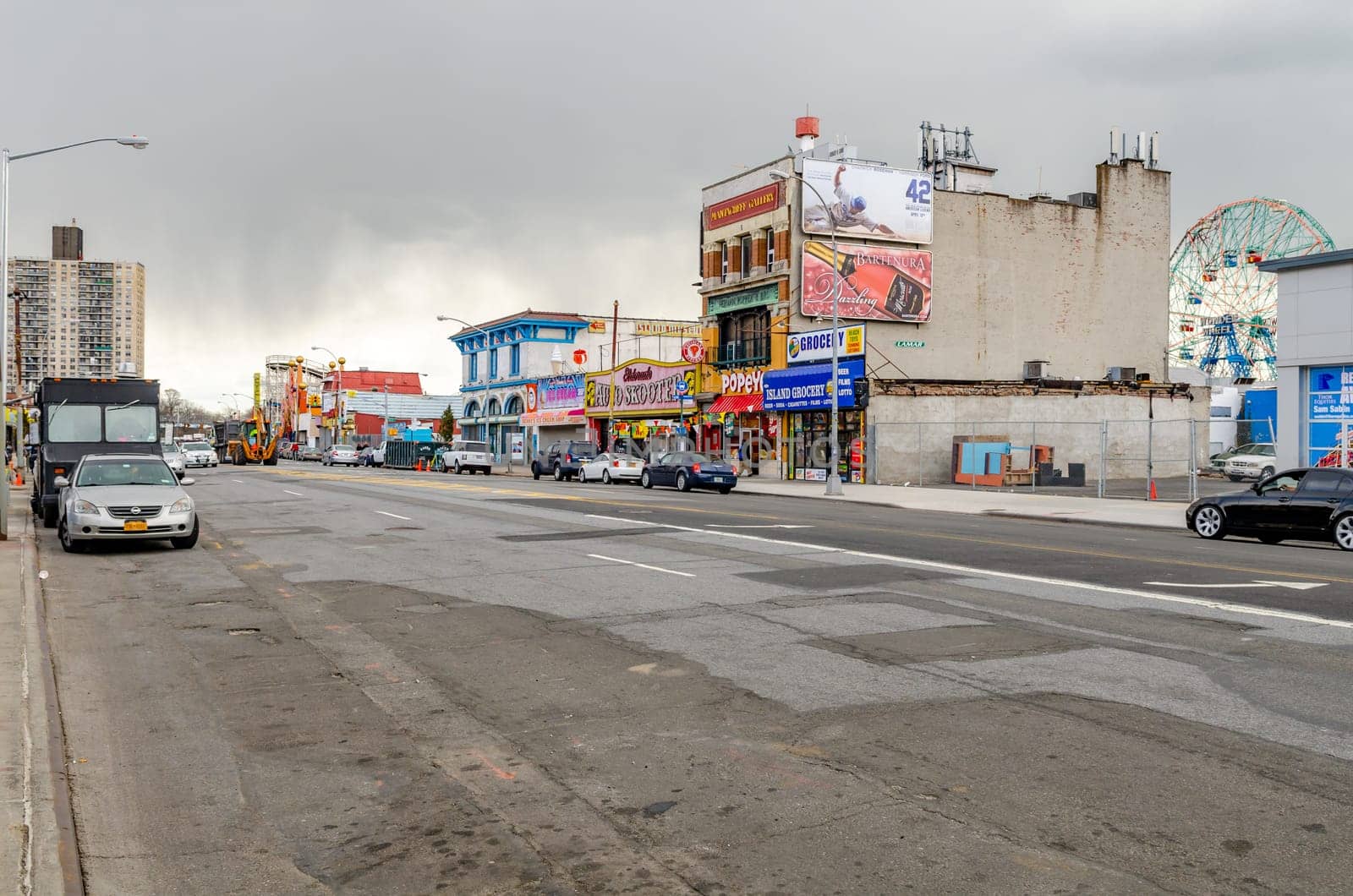 City Street with retail stores in Coney Island, ferris wheel behind the stores, cars parked next to the street, New York City during winter with overcast, horizontal