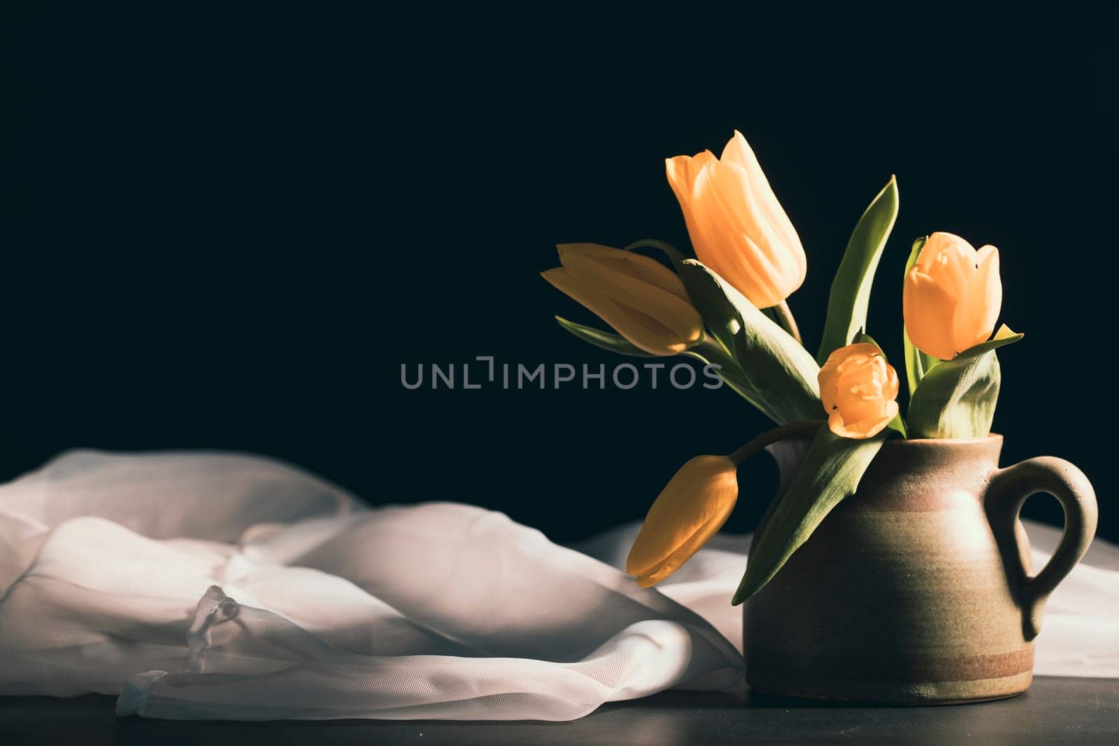 Still life with bouquet of yellow tulip flowers in old vintage jug, in beam of light on dark background.Art photography.