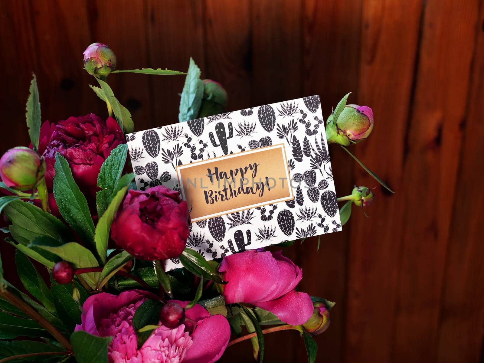 Close-up of pink peonies flowers and vase on wooden background with Happy Birthday card. The photo was taken with natural daylight.