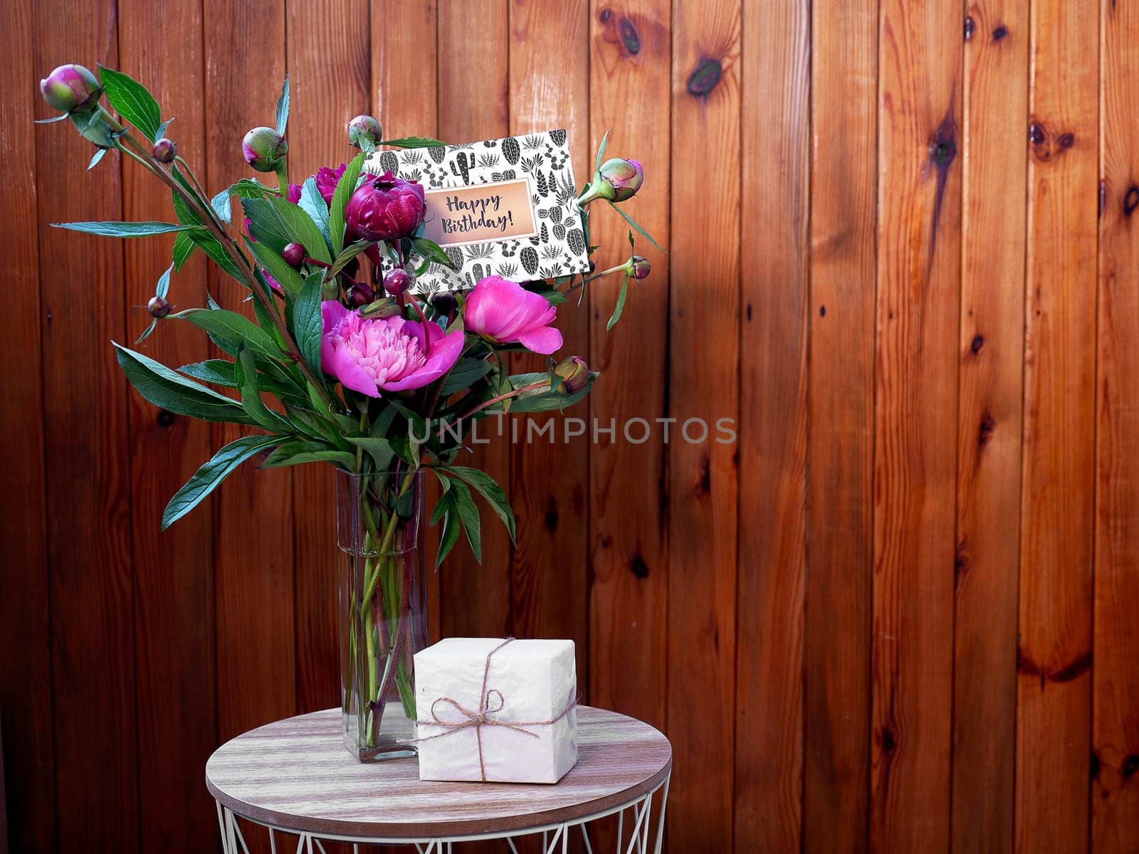 Pink flowers peonies in a vase on a wooden background with a small white gift and a happy birthday card. The photo was taken with natural daylight.