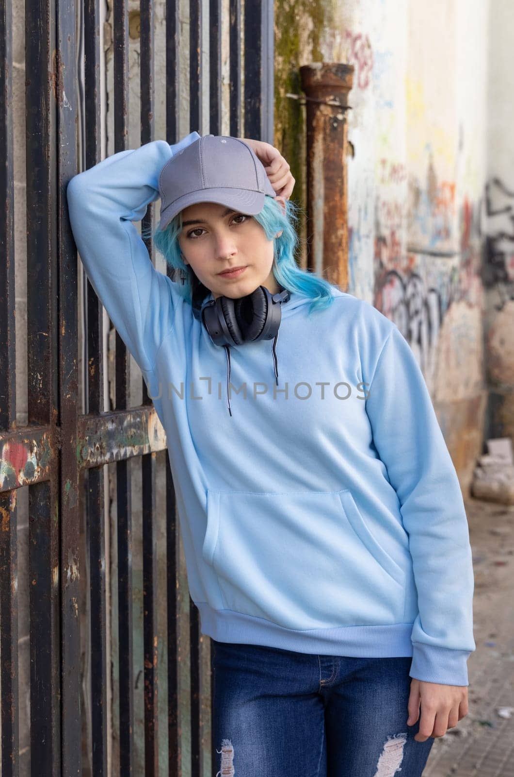 Teenager wearing light blue oversize hoodie and baseball cap with hand over head. Blue haired teen girl with wireless headphones stays outdoor near a gate. Clothing and baseball cap mockup