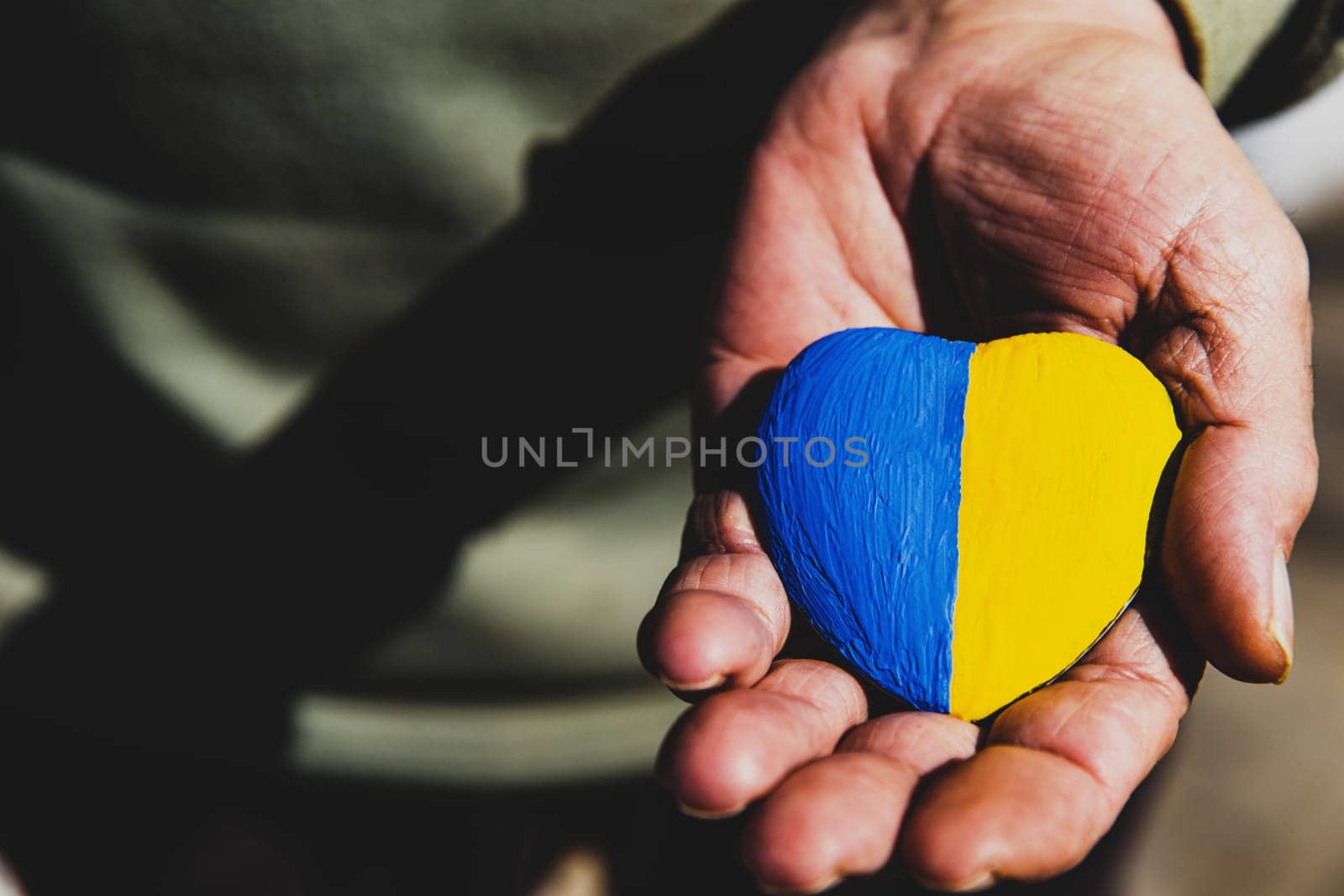 Senior man or soldier hands holding heart shape stone painted with Ukraine national flag colors. Pray for Ukraine, stop the war, save Ukraine people and children. No invasion, occupation and terrorism