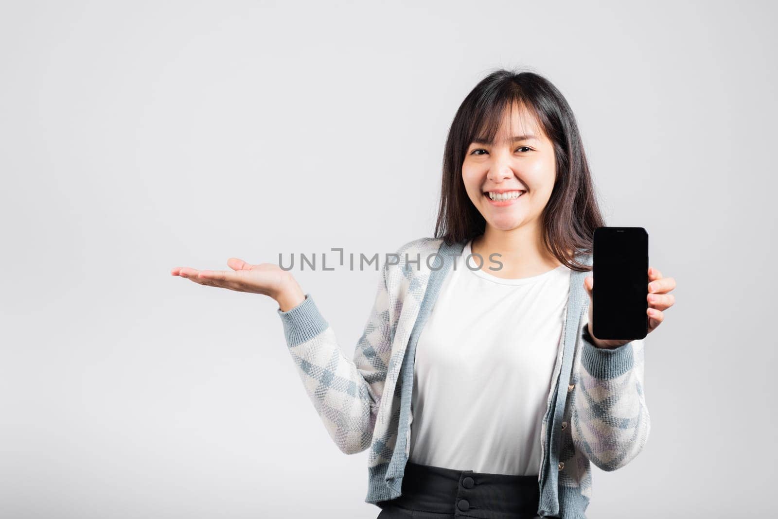 Woman excited holding mobile phone blank screen presenting product with palm of hand at empty studio shot isolated white background, happy young female smiling gesture show something product