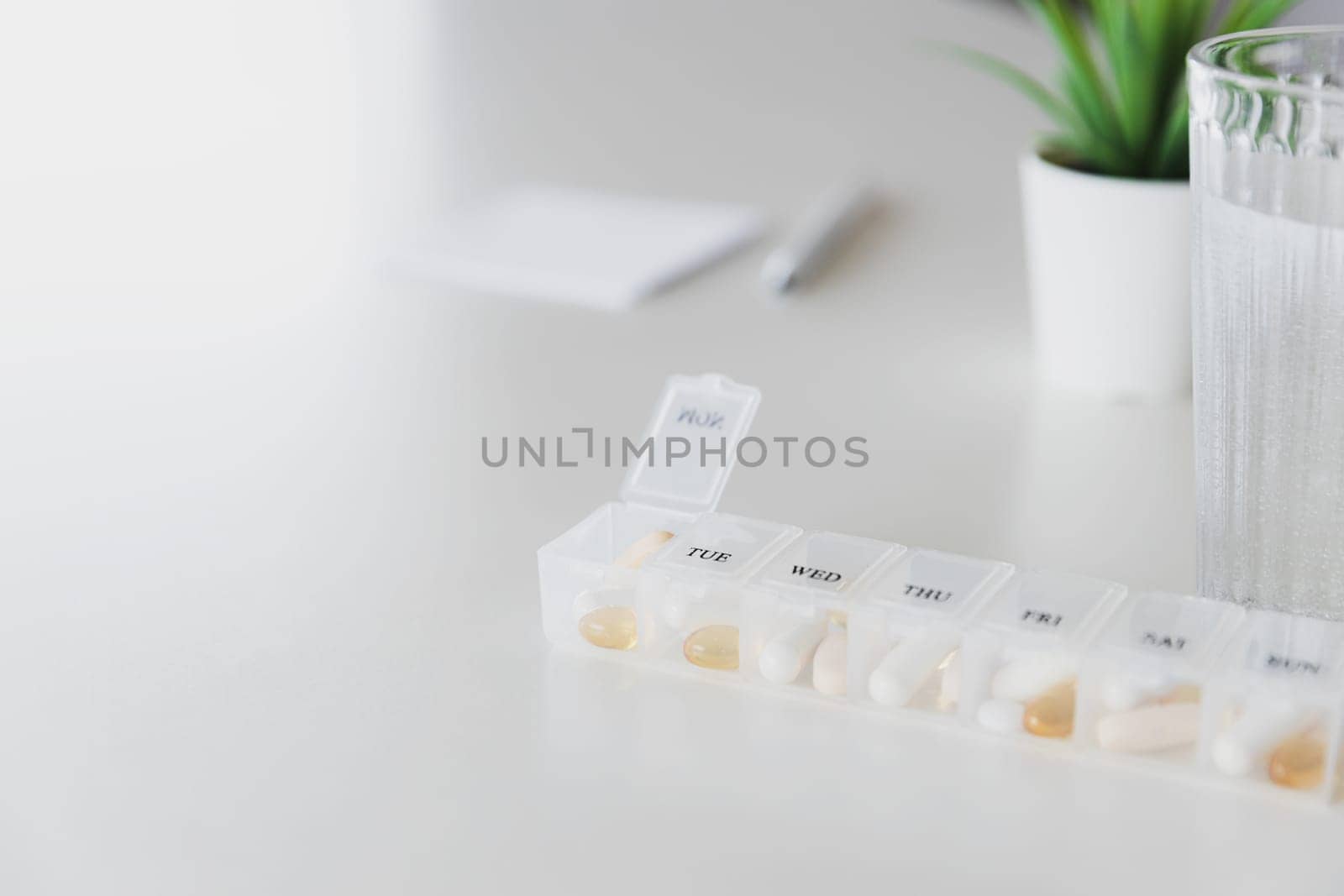 Closeup of medical pill box with doses of tablets for daily take a medicine with white, yellow drugs and capsules used for treatment, cure the disease. Glass cup of water on table, gray background.