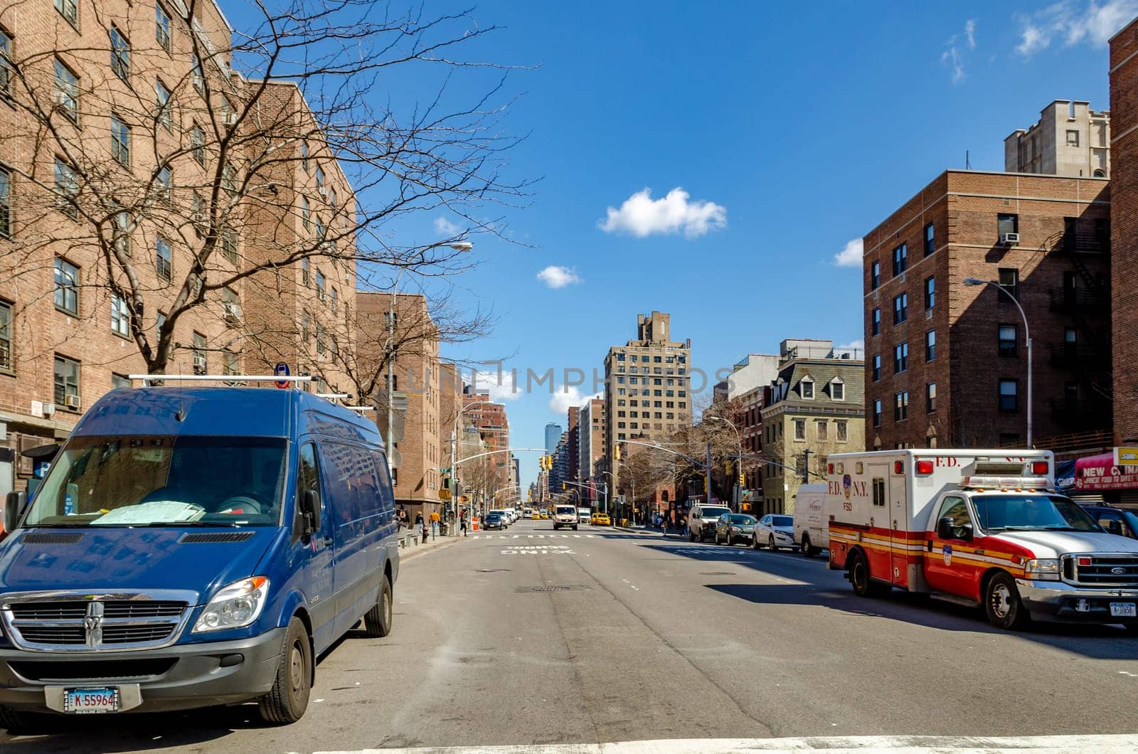 City Street in Chelsea with cars parked next to the street and traffic, emergency car passing by, clear sky, New York City during sunny winter day, horizontal