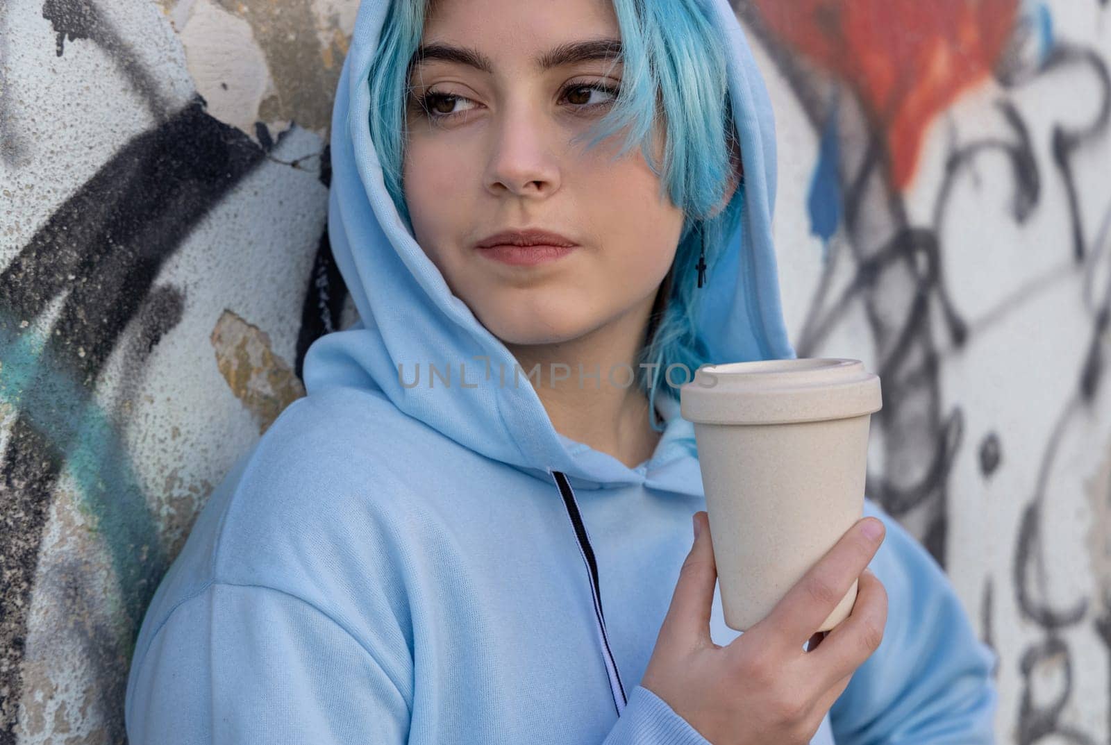 Teenage girl in light blue oversize hoodie with coffee to go and looking aside. Blue haired teen girl staying outdoors near graffiti wall. Cup mockup. Hipster and adolescence concept