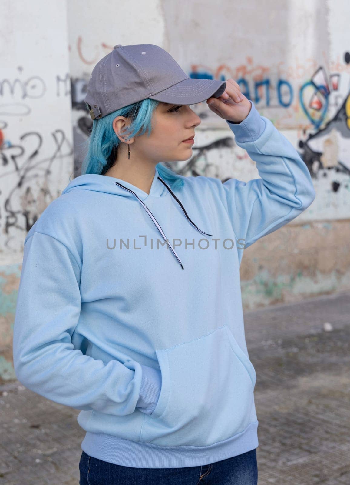 Blue haired Teenage girl in blue hoodie and baseball cap touching her cap and looking right. Blue haired teen girl stays outdoors against graffiti wall. Clothing and cap mockup