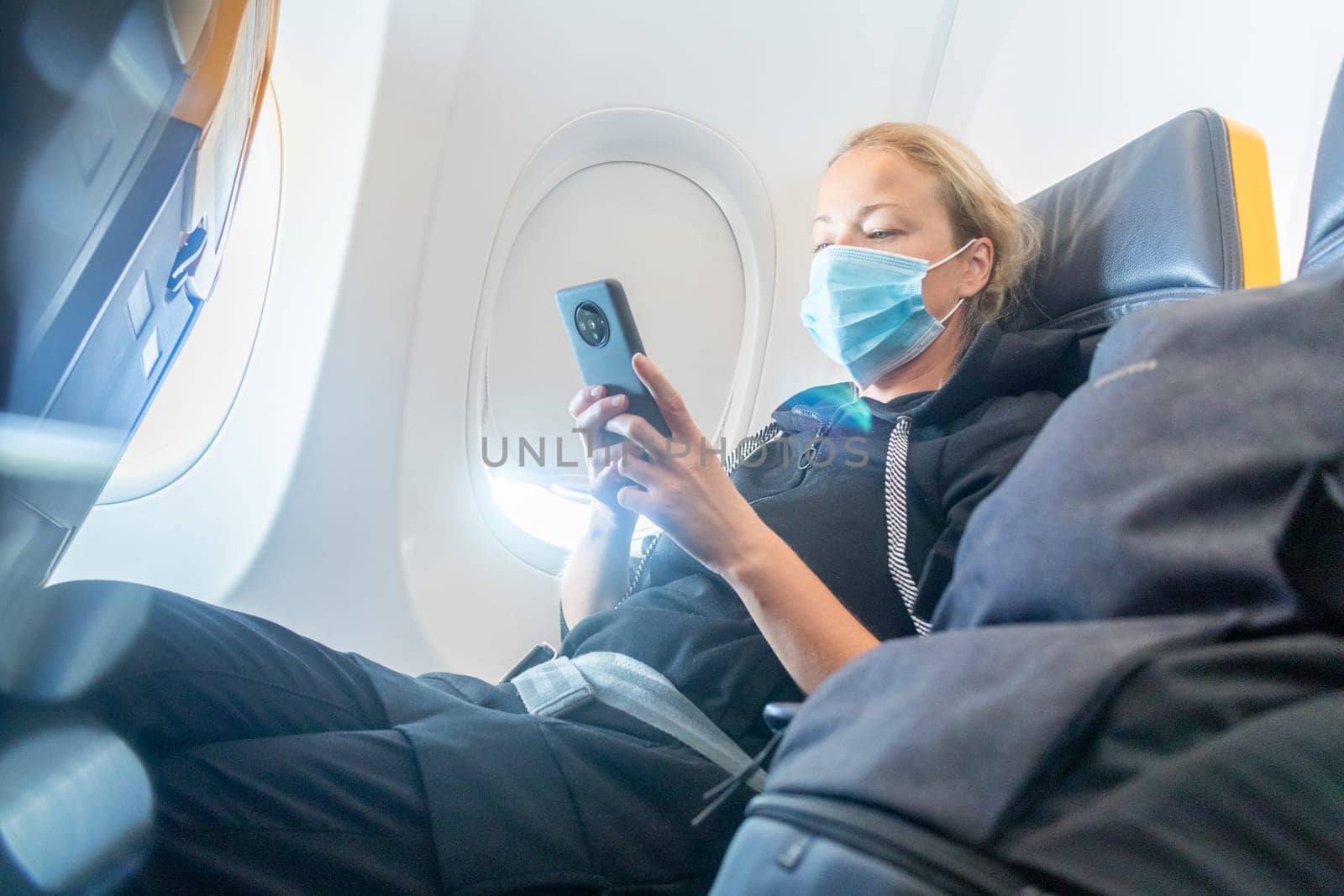 A young woman wearing face mask, using smart phone while traveling on airplane. New normal travel after covid-19 pandemic concept