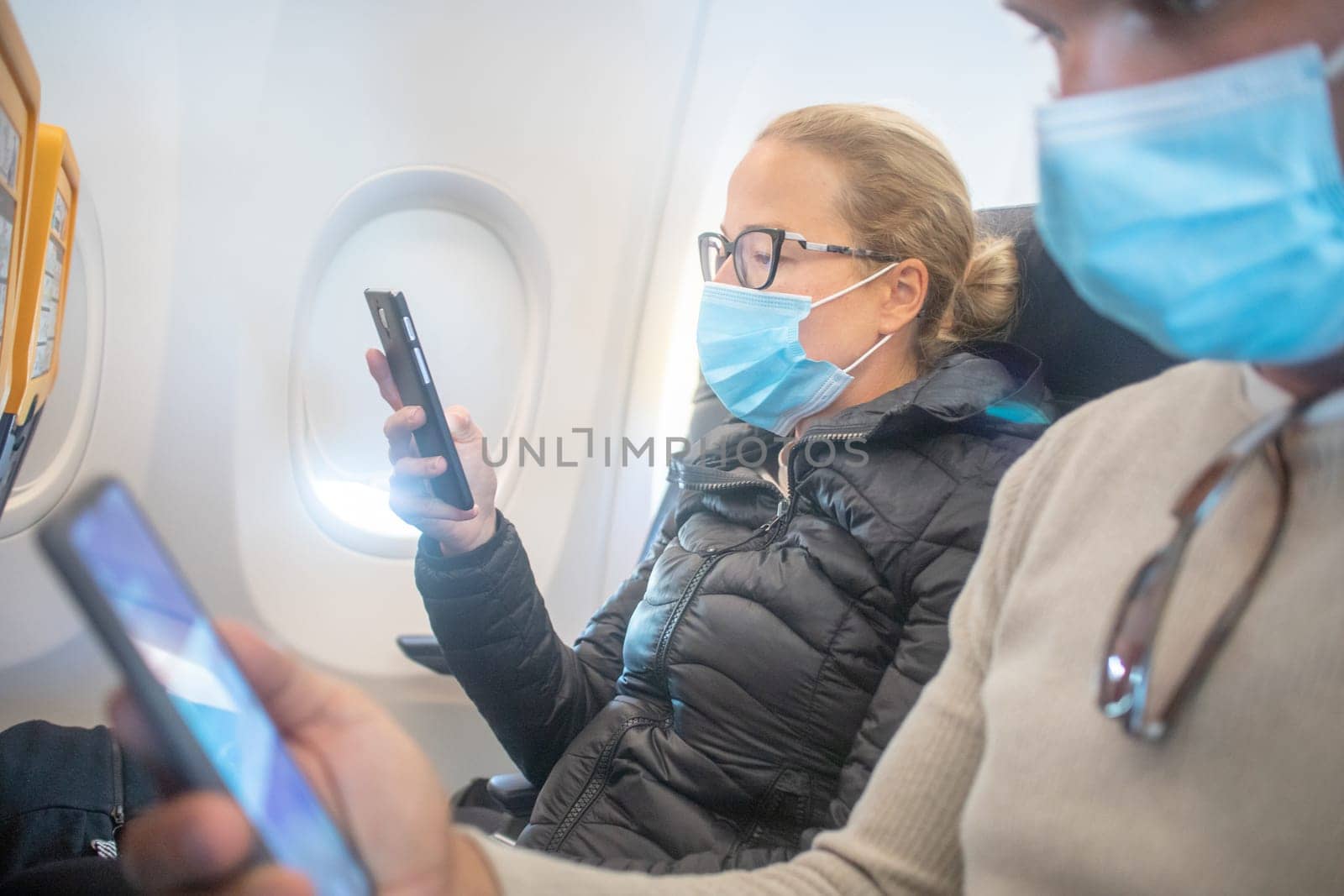 A young couple wearing face mask, using smart phone while traveling on airplane. New normal travel after covid-19 pandemic concept