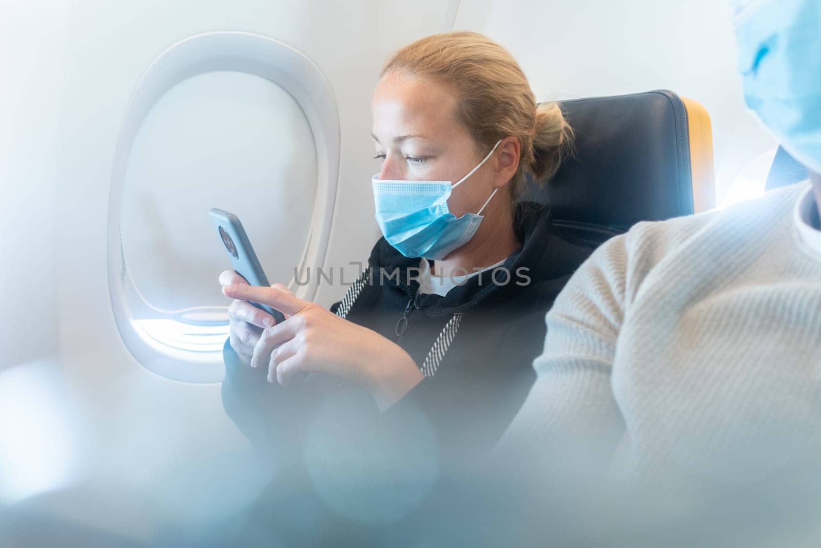 A young woman wearing face mask, using smart phone while traveling on airplane. New normal travel after covid-19 pandemic concept