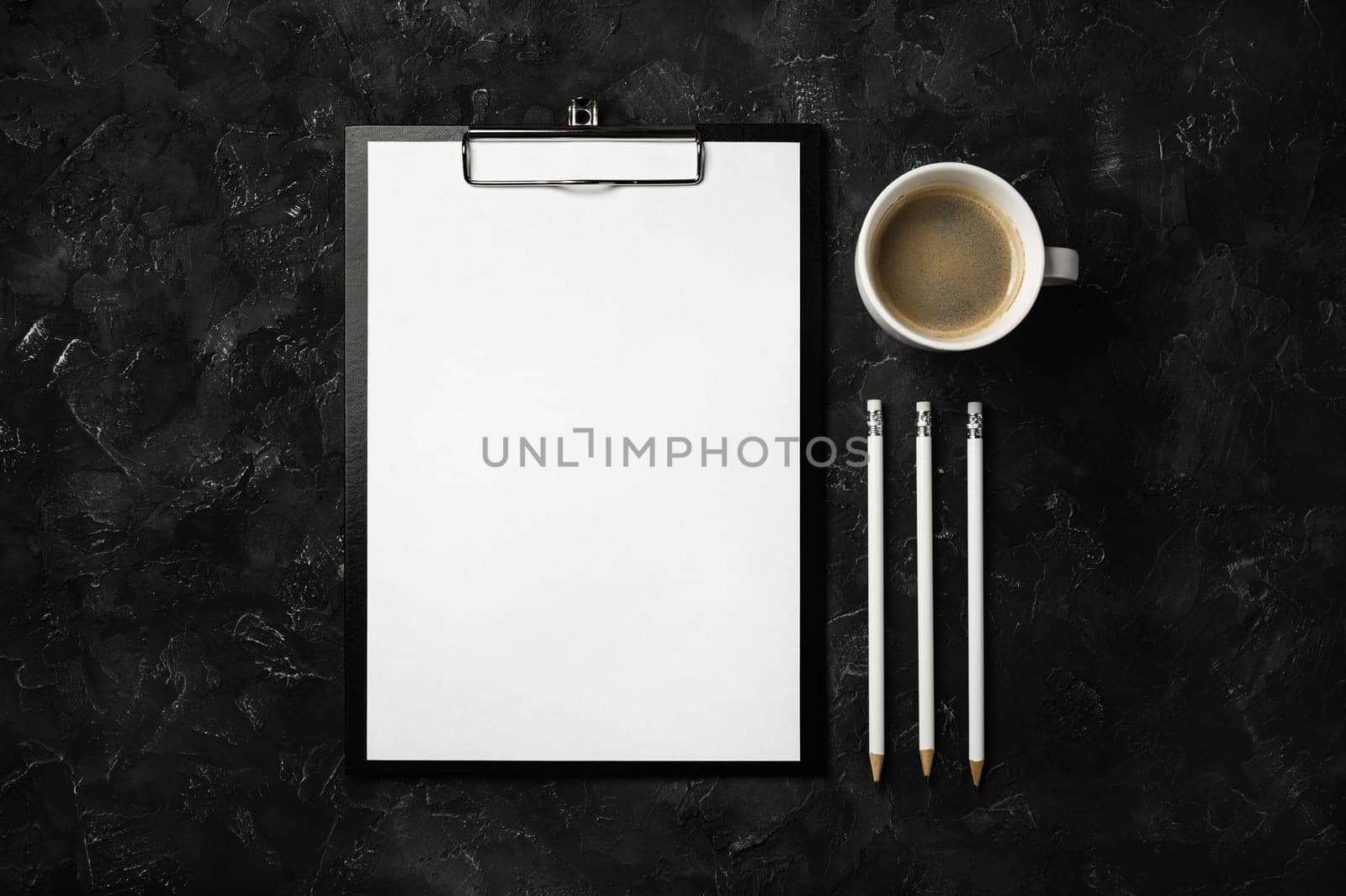 Photo of clipboard with blank paper, coffee cup and pencils on black plaster background. Office desk concept.