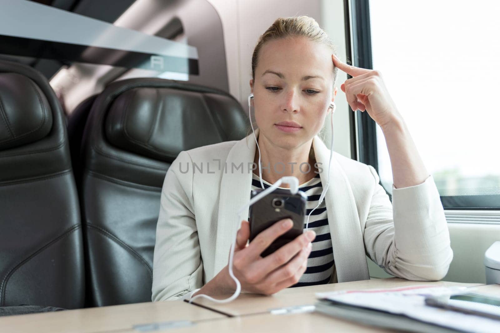 Caucasian businesswoman communicating on cellphone using headphone set while traveling by train in business class seat.