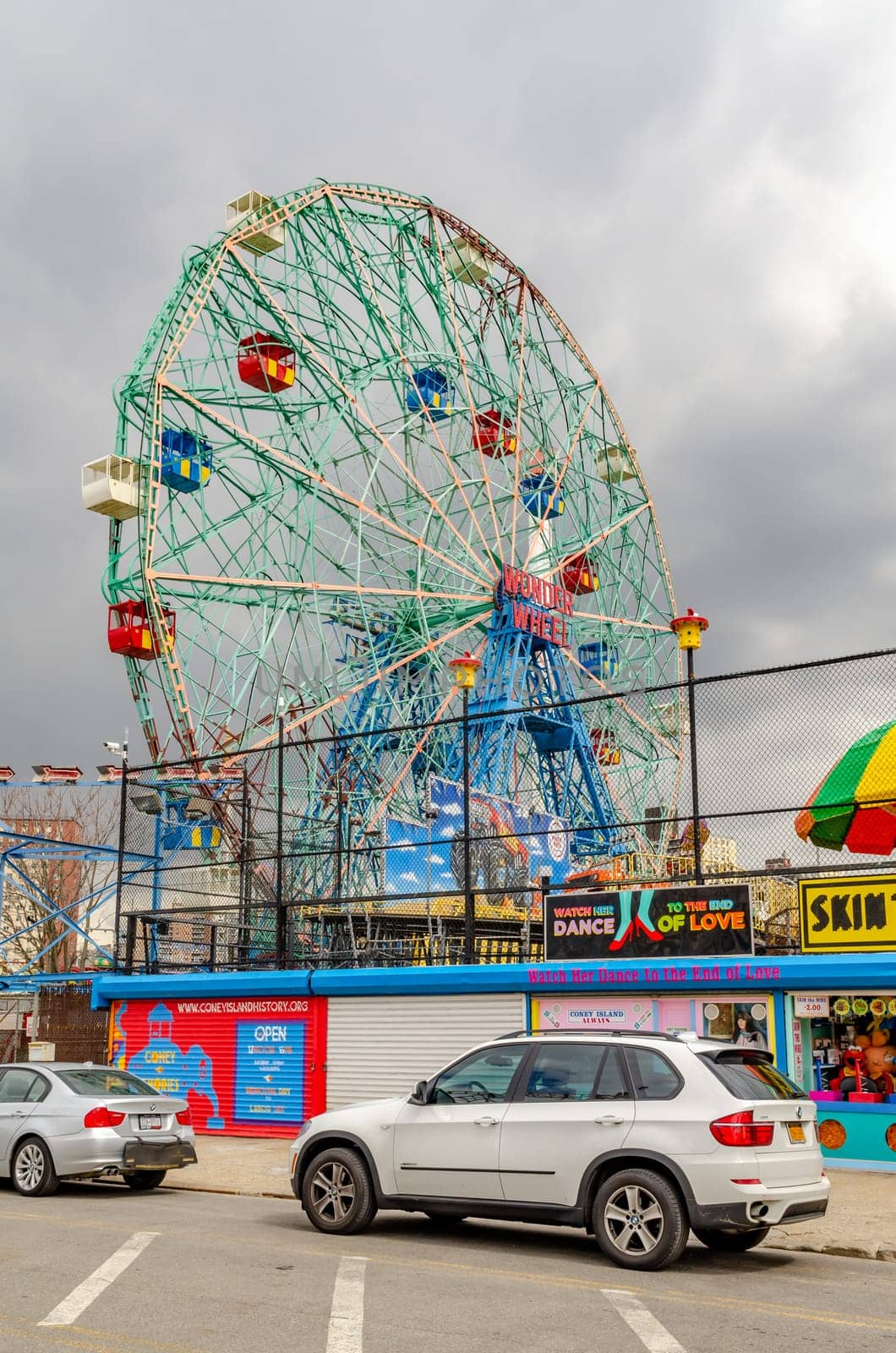 Wonder Wheel Ferris Wheel at Luna Park Amusement Park with stores and vending machines in front, Coney island, Brooklyn, New York City during winter day with cloudy sky, Cars parked next to the street in front, vertical