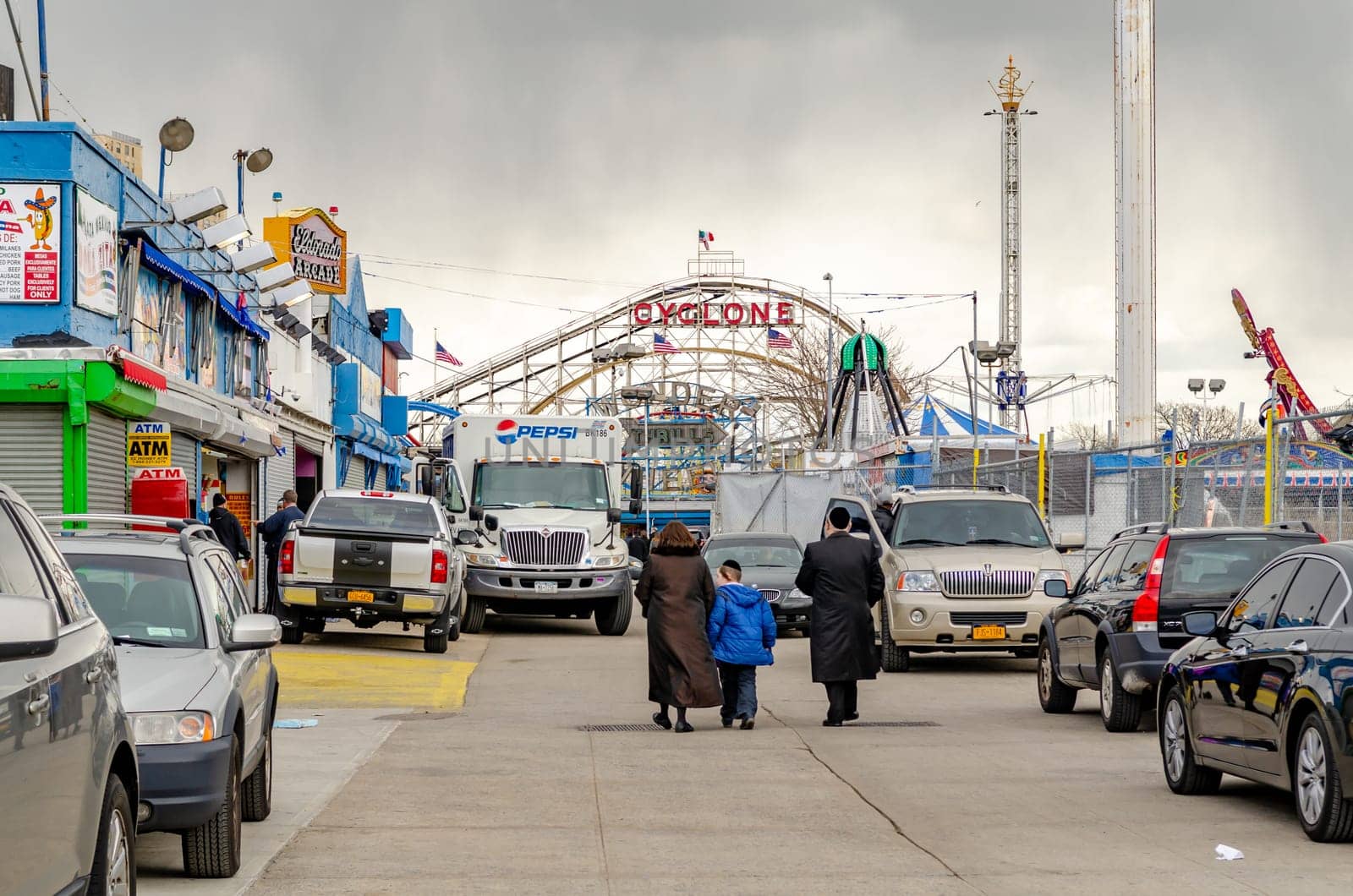 Jewish National Day at Luna Park Amusement Park, Coney island, Brooklyn, New York City during winter day with cloudy sky, Family walks in the direction of the Park, lots of cars parked next to the street, horizontal