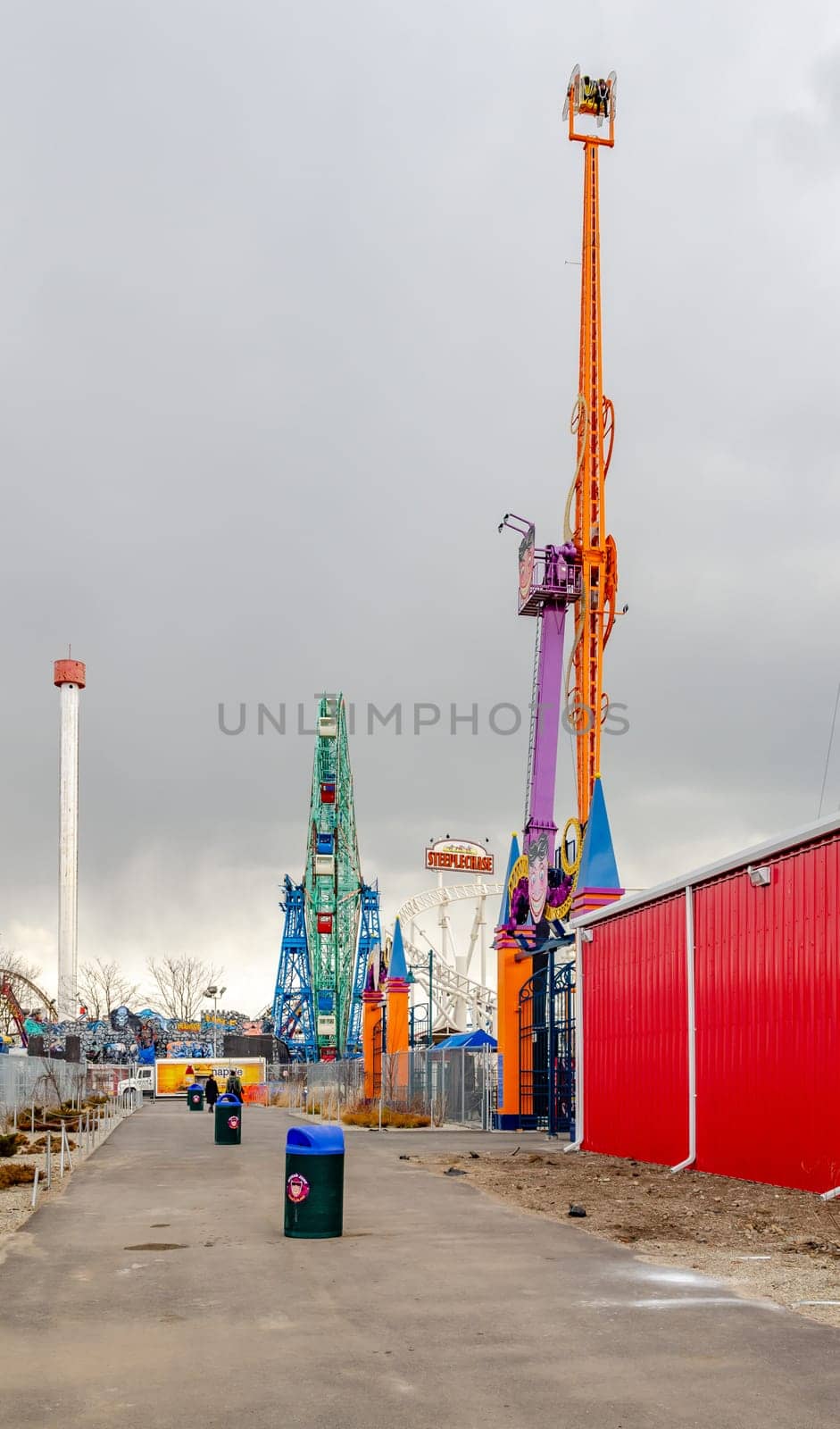 Wonder Wheel, Ferris Wheel and other Rides at Luna Park Amusement Park, Coney island, Brooklyn, New York City during winter day with cloudy sky, Trash cans standing on a footpath in front, vertical