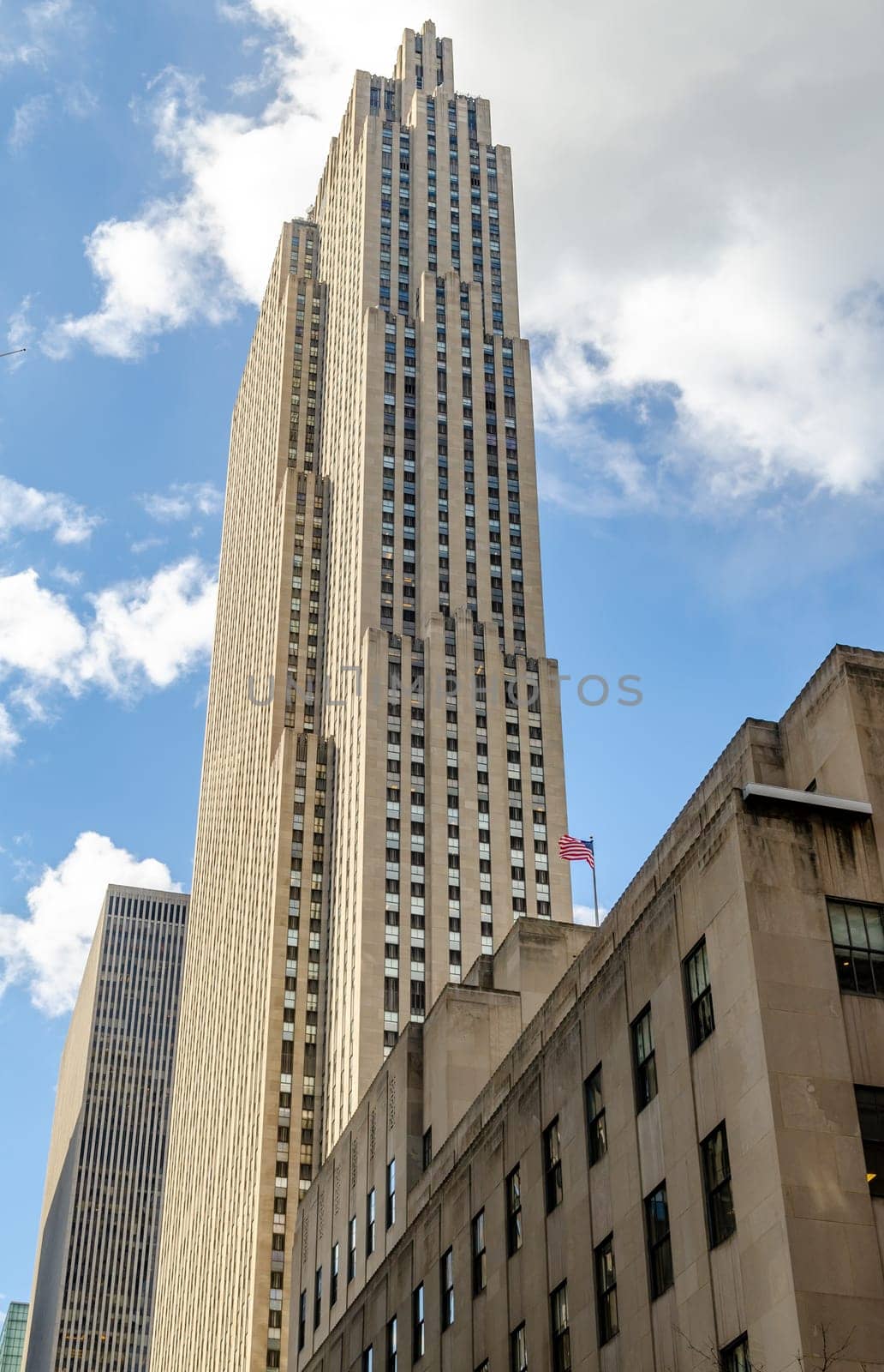 Rockefeller Center Skyscraper, view from the side, low angle during winter day with cloud sky, American flag in front of the Building, vertical