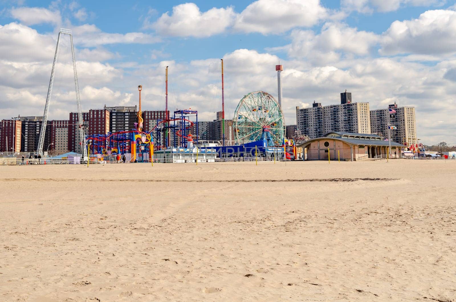 Coney island, Brooklyn Luna Park Amusement Park with Rollercoaster, Ferris Wheel and Tower, Beach in front during sunny winter day with cloudy sky, no people, view from the side, New York City, Residential Buildings in the Background, horizontal