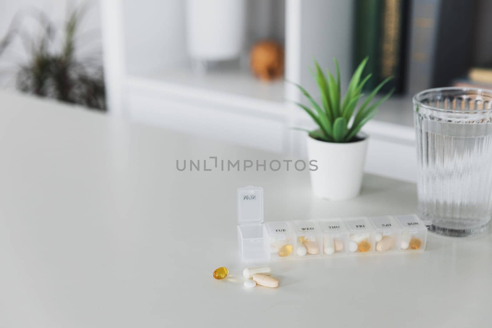 Closeup of medical pill box with doses of tablets for daily take a medicine for treatment, cure the disease. Glass cup of water on table, gray background. Green flowerpot or plant. Helathy lifestyle