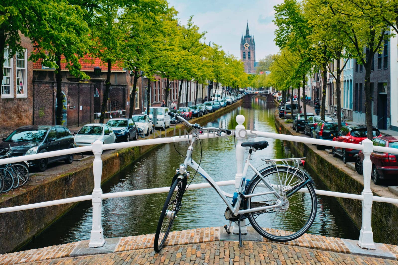 Picturesque Holland cityscape view - bicycle on bridge and canal with cars parked along and Oude Kerk old church in street of Delft, Netherlands