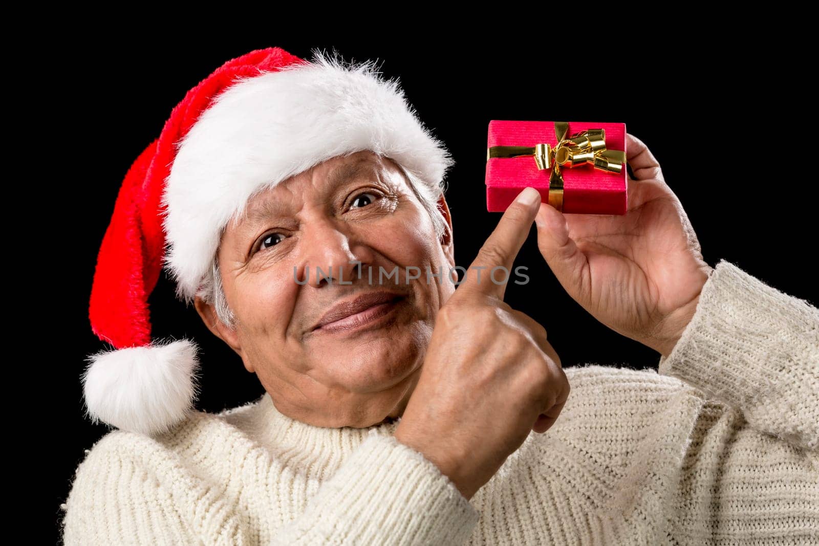 Pensive aged man in warm pullover and red Santa Claus cap. He is pointing his right index finger at a small, red, wrapped gift held on eye-level in his left hand. Isolated on black. Gift-giving theme.
